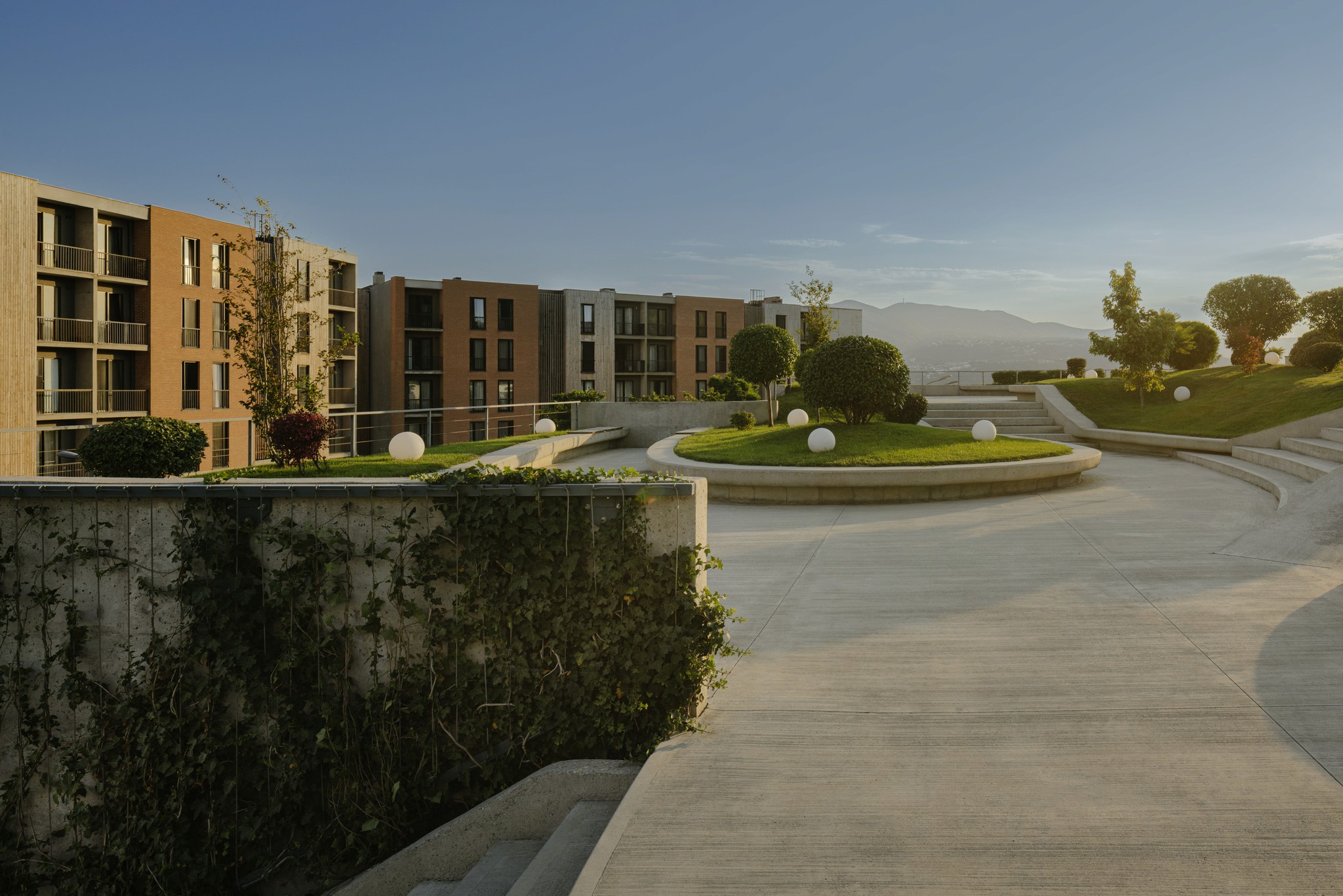 A modern building with greenery, pathways, and mountains in the background under a clear blue sky.