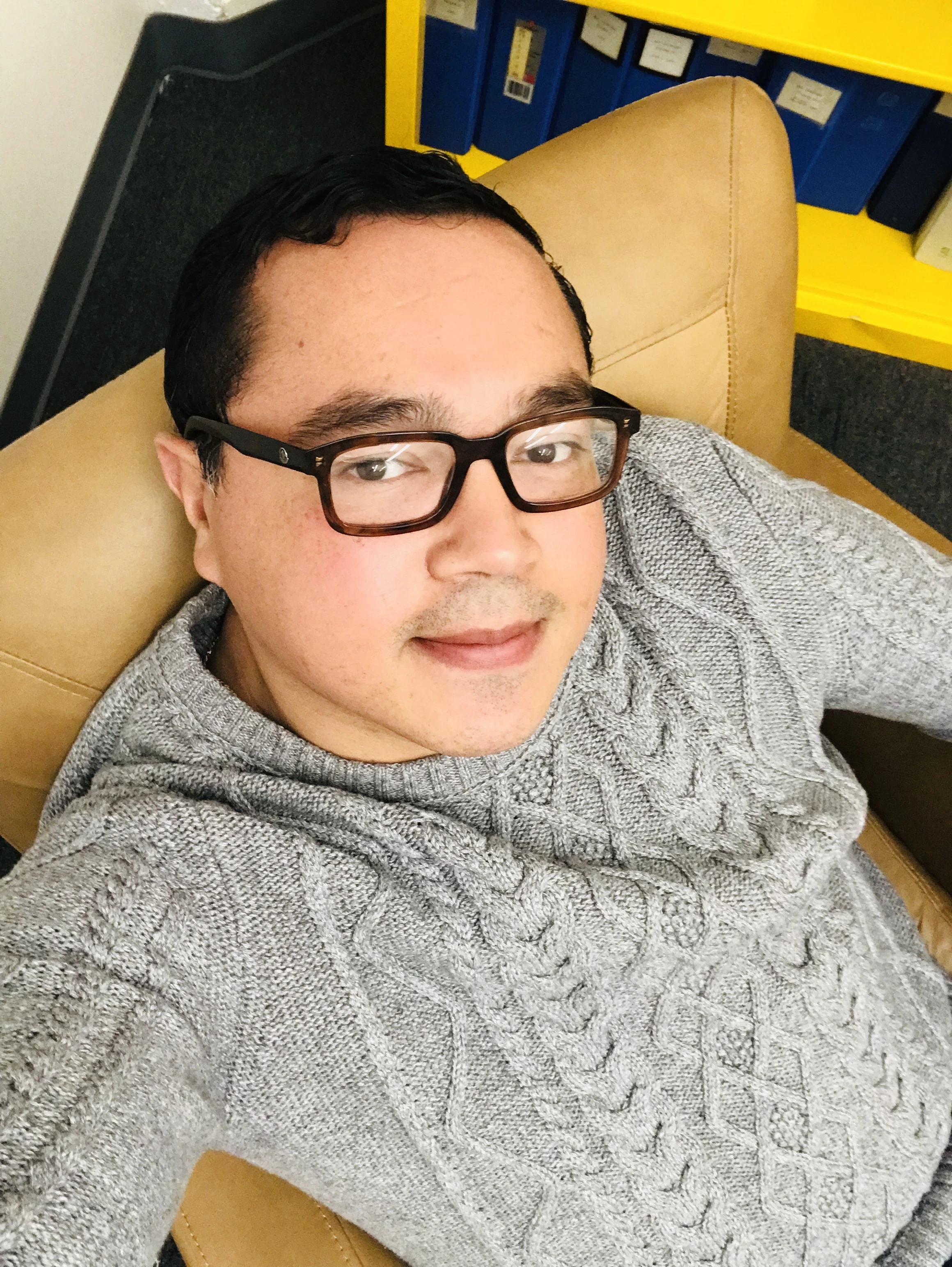 Uma pessoa usando óculos e um suéter cinza de tricô trançado está sentada em uma cadeira de couro bege, com uma estante amarela e pastas azuis ao fundo.