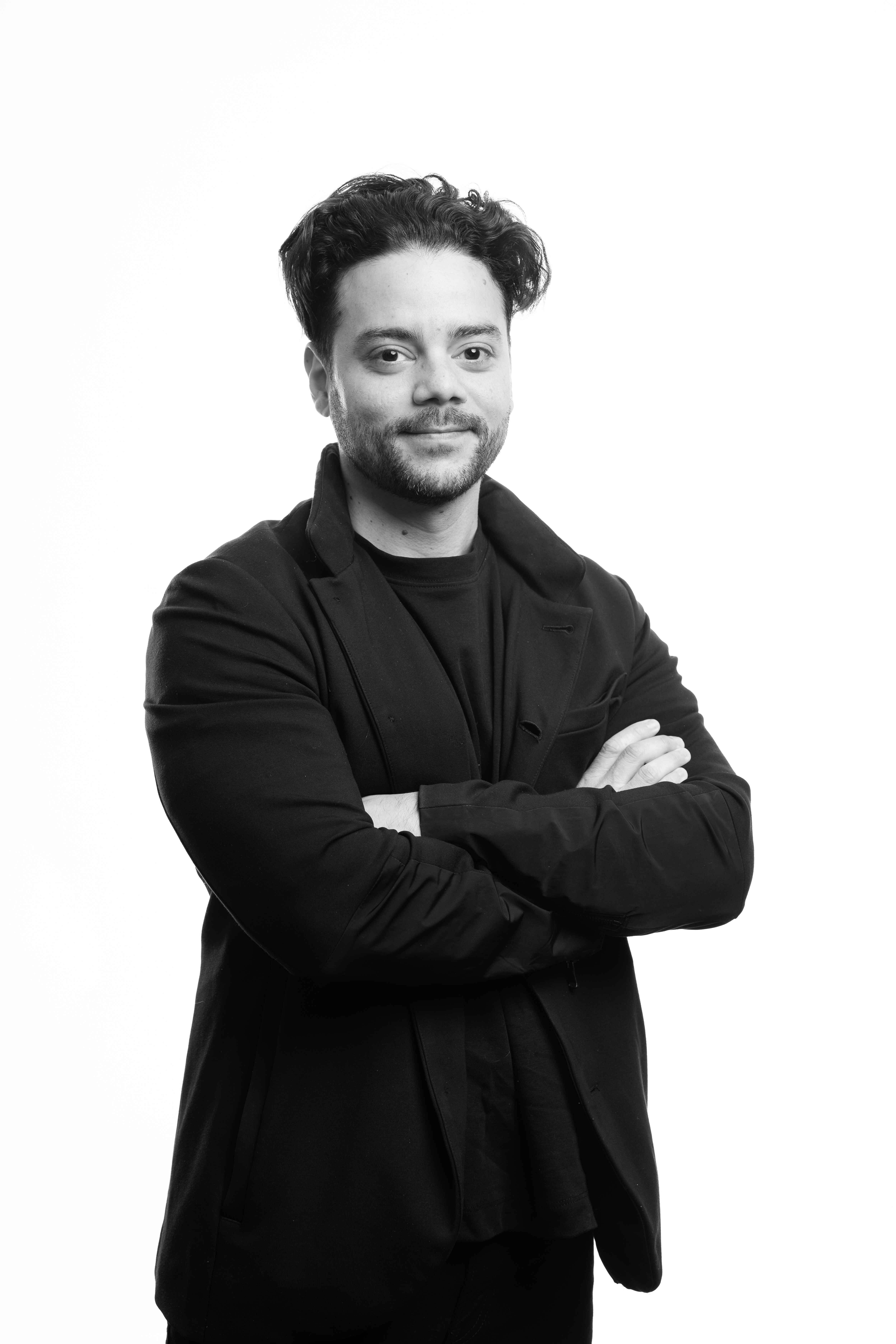 Black and white portrait of a man with curly hair, wearing a black jacket, standing confidently with crossed arms against a plain background.