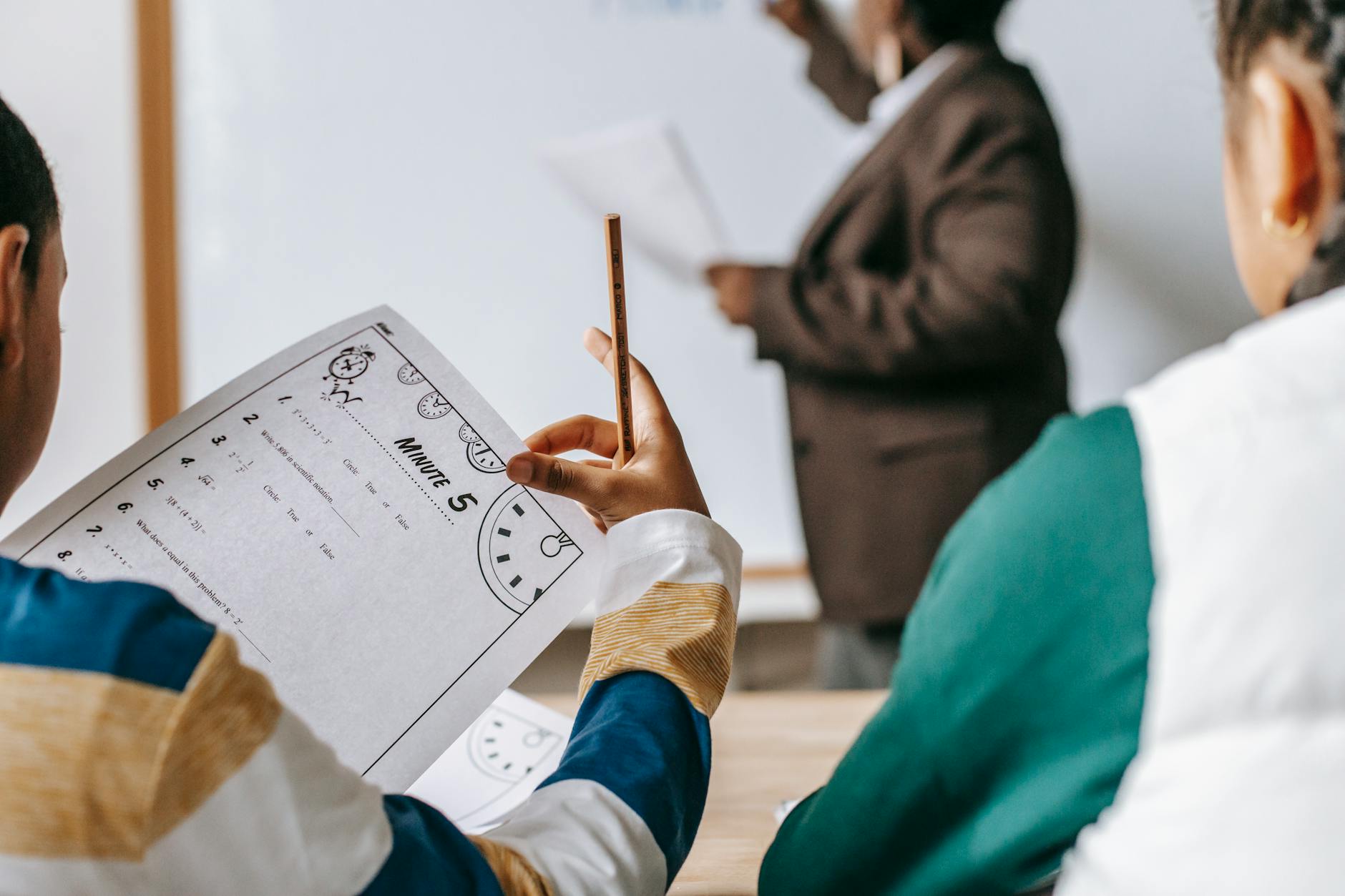 A smiling educator handing printed worksheets to a diverse group of elementary students in a bright classroom.