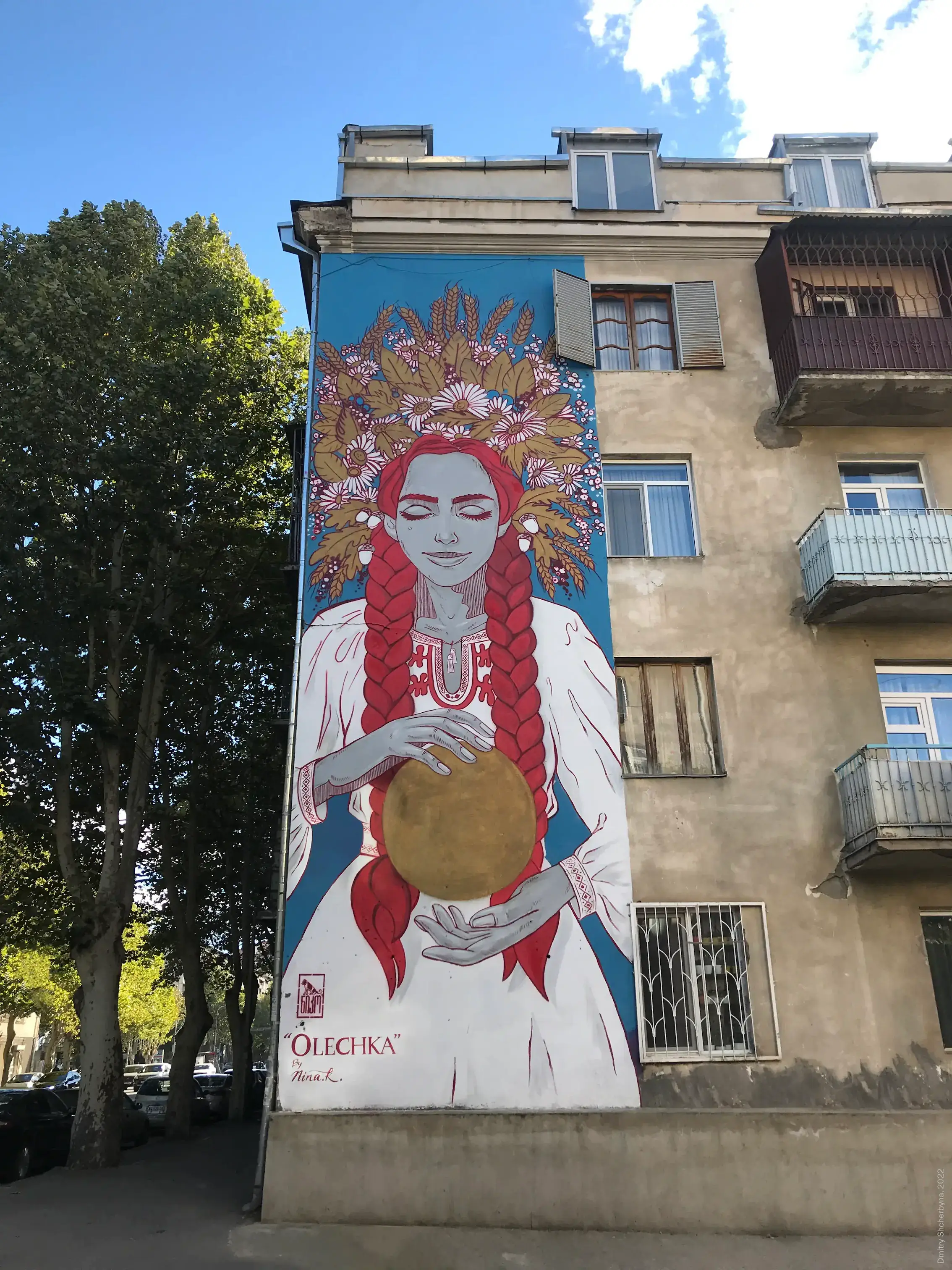A mural on the side of an apartment building shows a serene woman with red braids, closed eyes, and an elaborate wreath of wheat, flowers, and leaves on her head. She is wearing a white embroidered blouse and holding a golden orb.