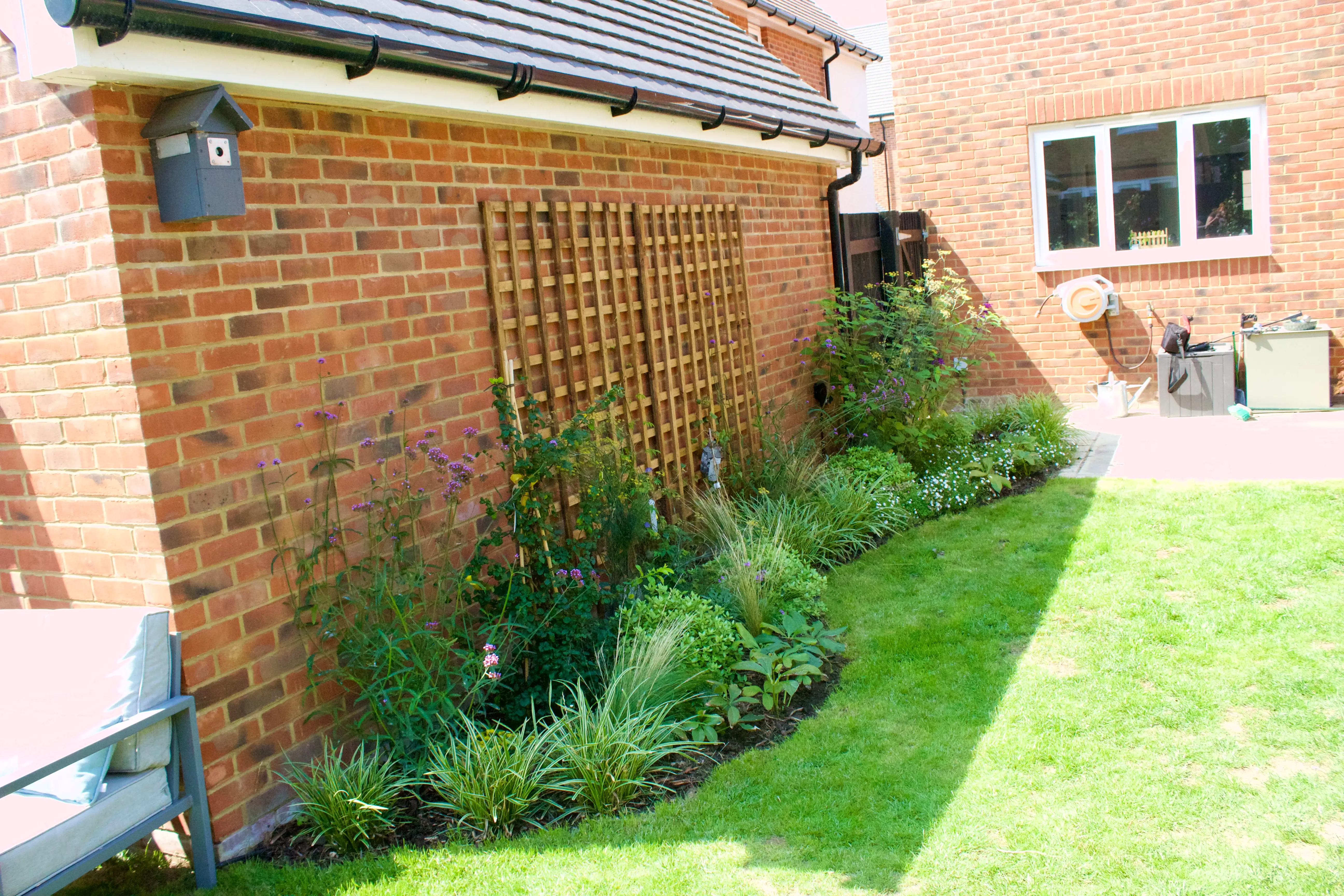 A brick wall of a house with greenery and grass along the side, partially shaded by the building.