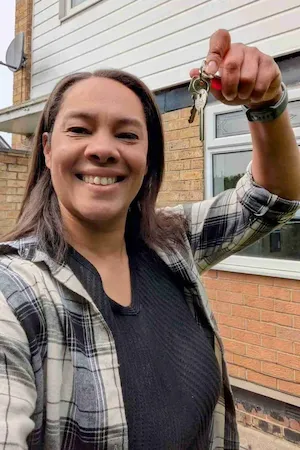 Woman in plaid flannel shirt smiling and holding up house keys in front of a residential property