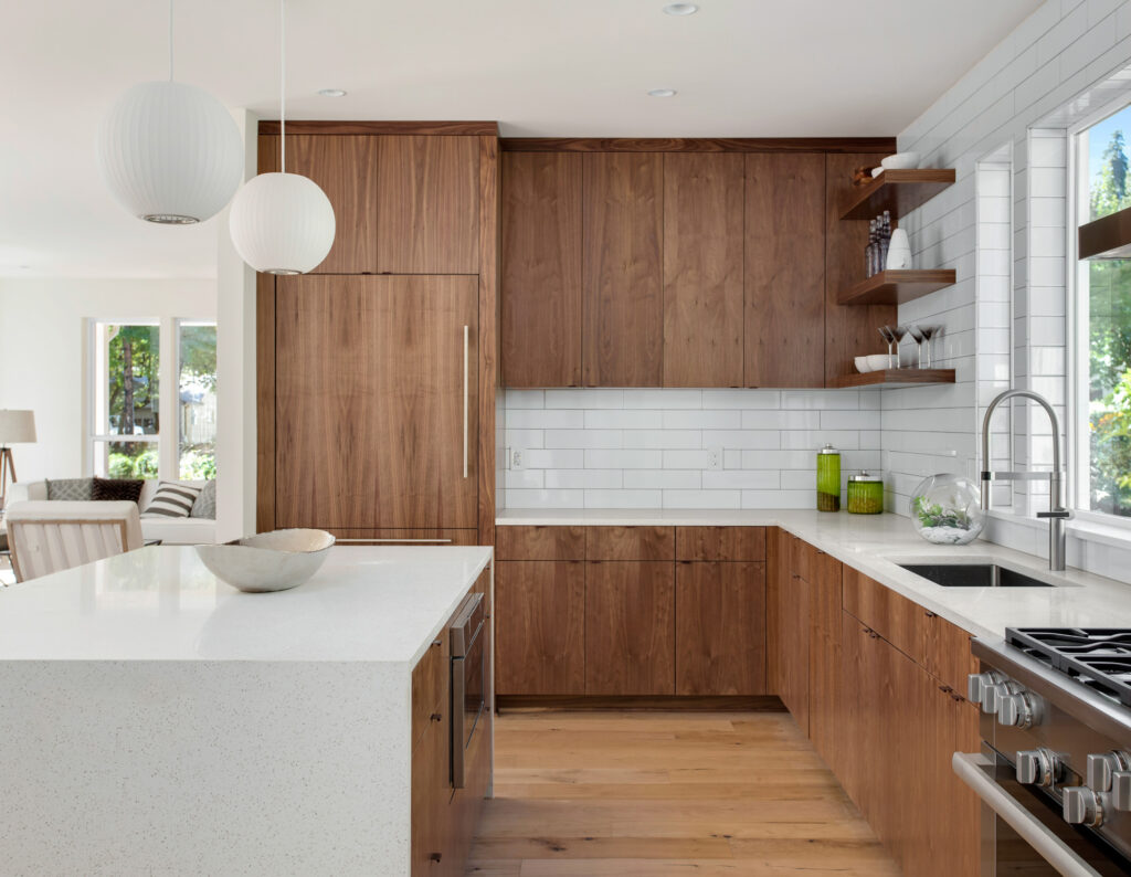 Modern kitchen with white walls and backsplash, natural wood cabinets, a white island with a waterfall countertop, and four metal barstools. Two round pendant lights hang above the island, and a stainless steel oven and hood are installed along the back wall with windows above the countertop.