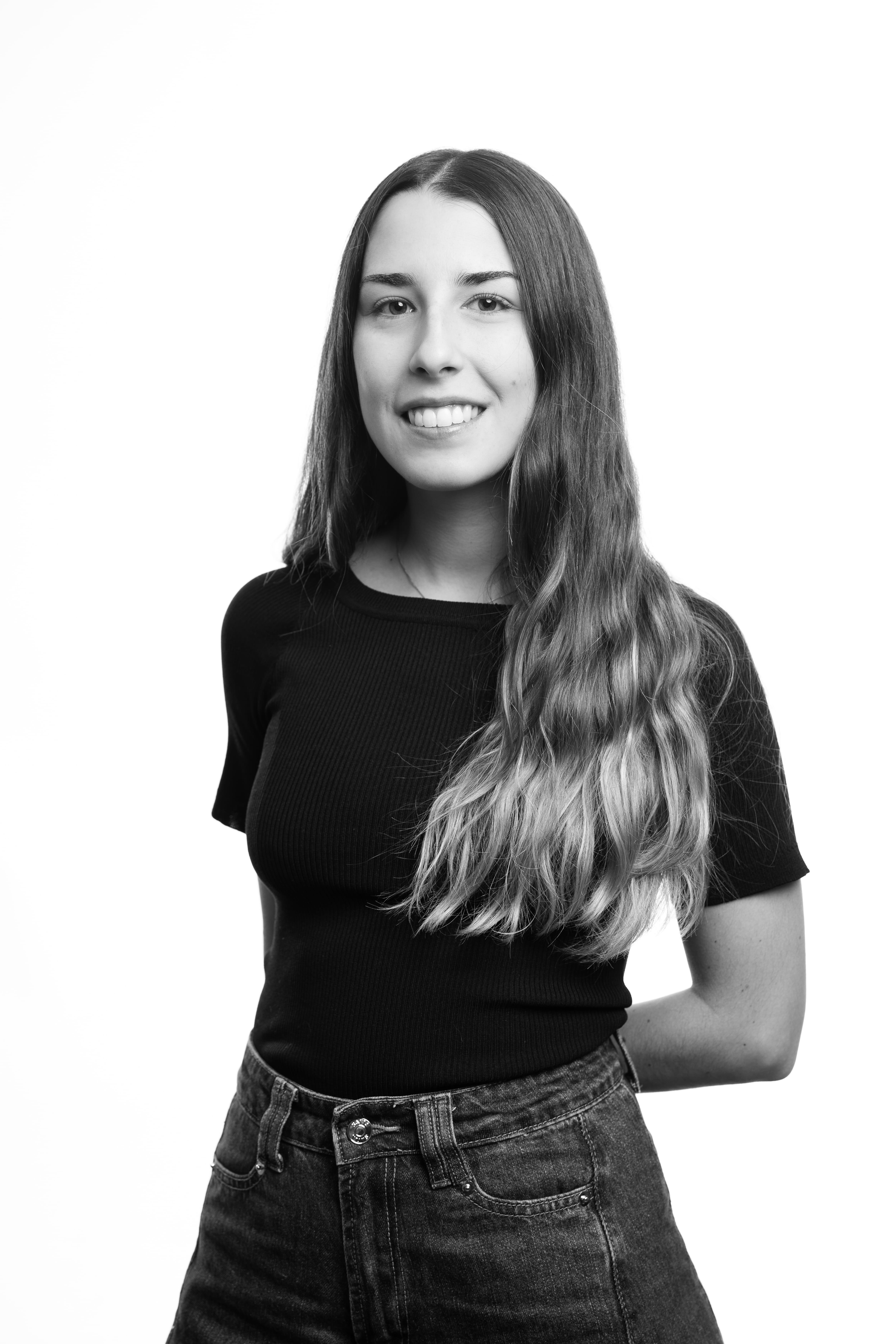 A woman with long hair smiles in a black and white portrait. She's wearing a black shirt and jeans, standing with her hands behind her back, conveying a relaxed and approachable mood.