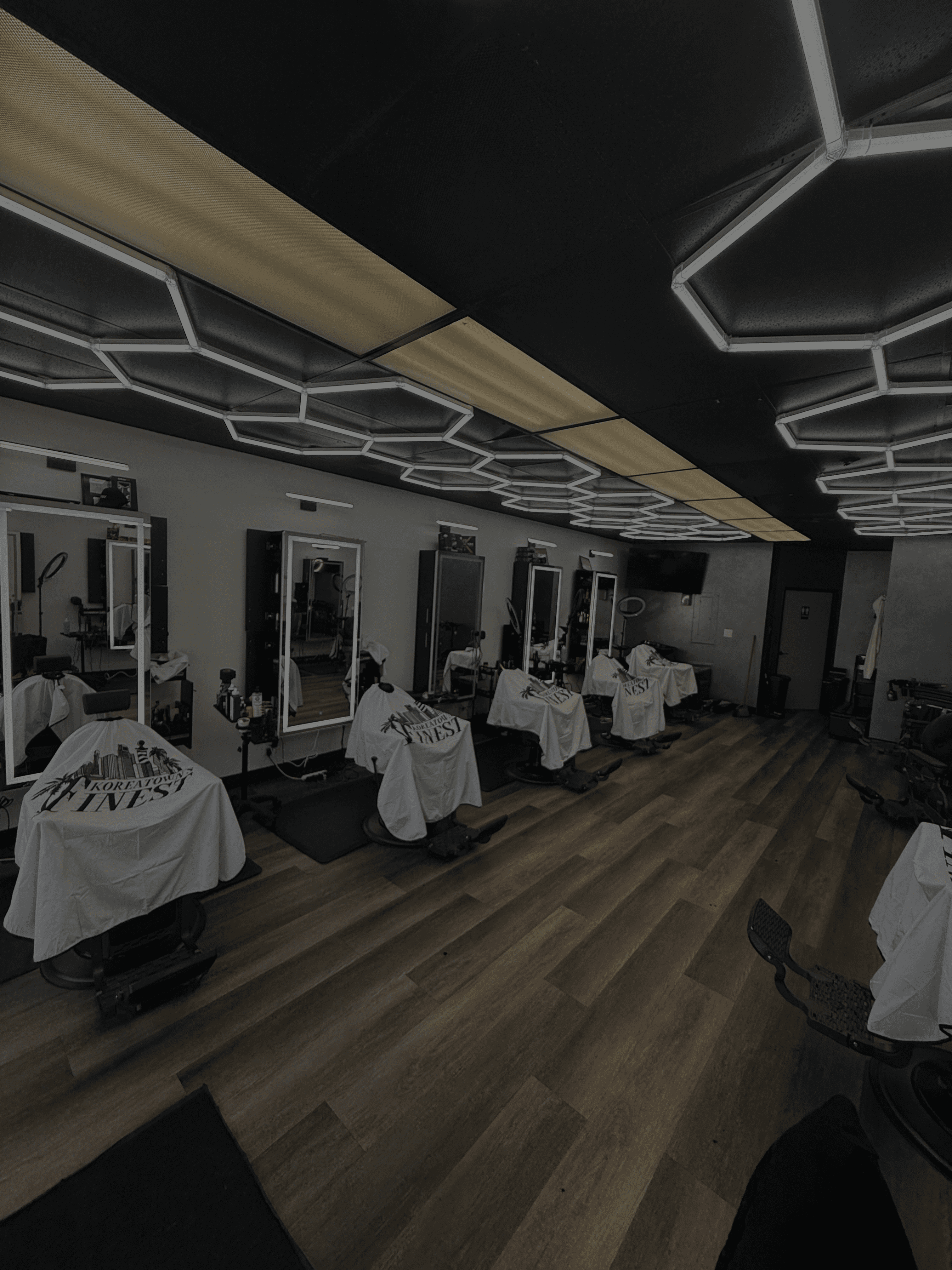 Modern barbershop interior with five barber chairs lined against a wall of mirrors, bright geometric ceiling lights, and wood-style flooring..
