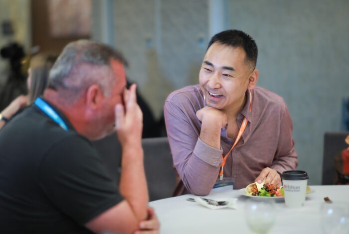 Two men sit at a round table, laughing and talking during a meal. One man holds his head while the other, smiling with his chin in hand, has a plate of food and a Stukent-branded cup in front of him.