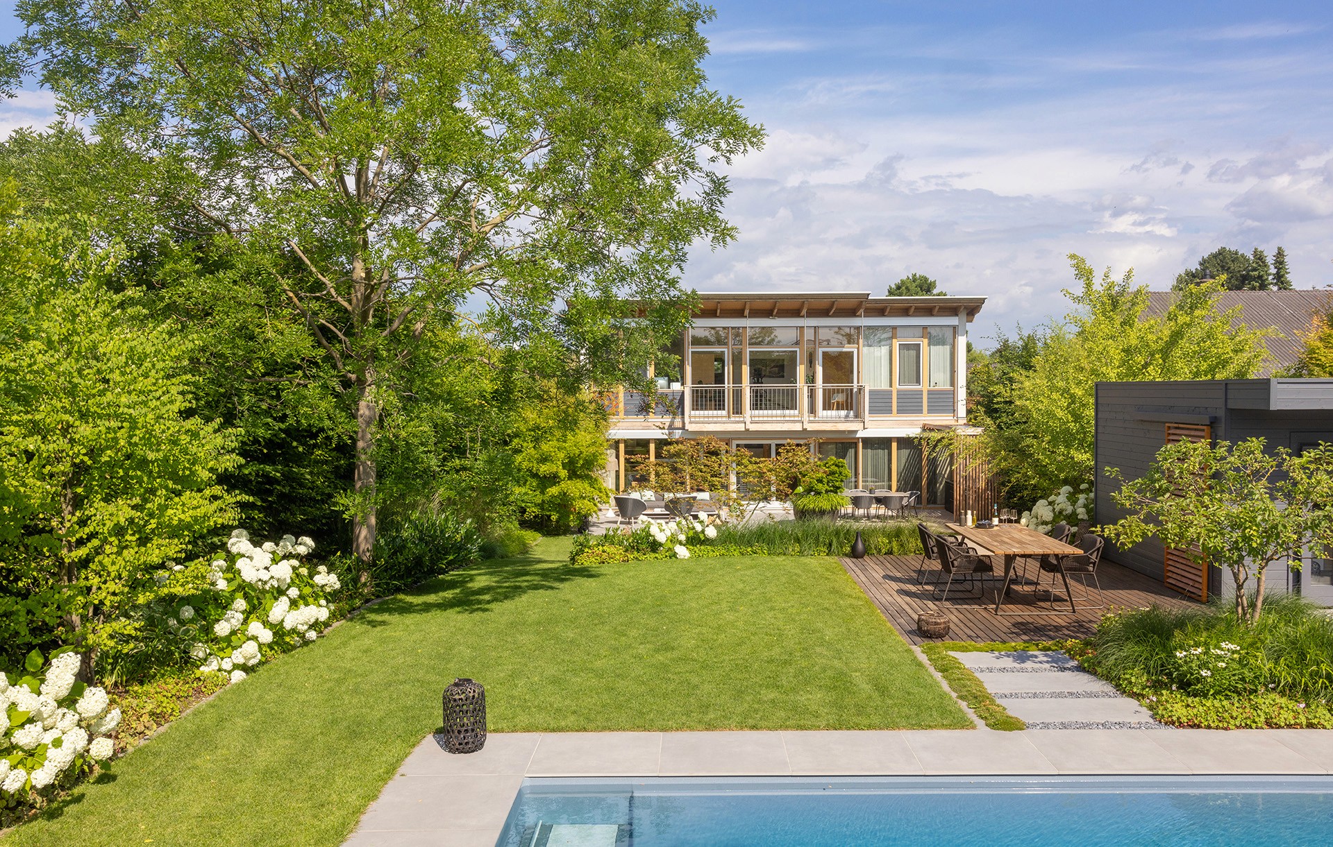 Moderner Garten mit Pool, Rasenfläche, großen Bäumen und Holzterrasse mit Essbereich.