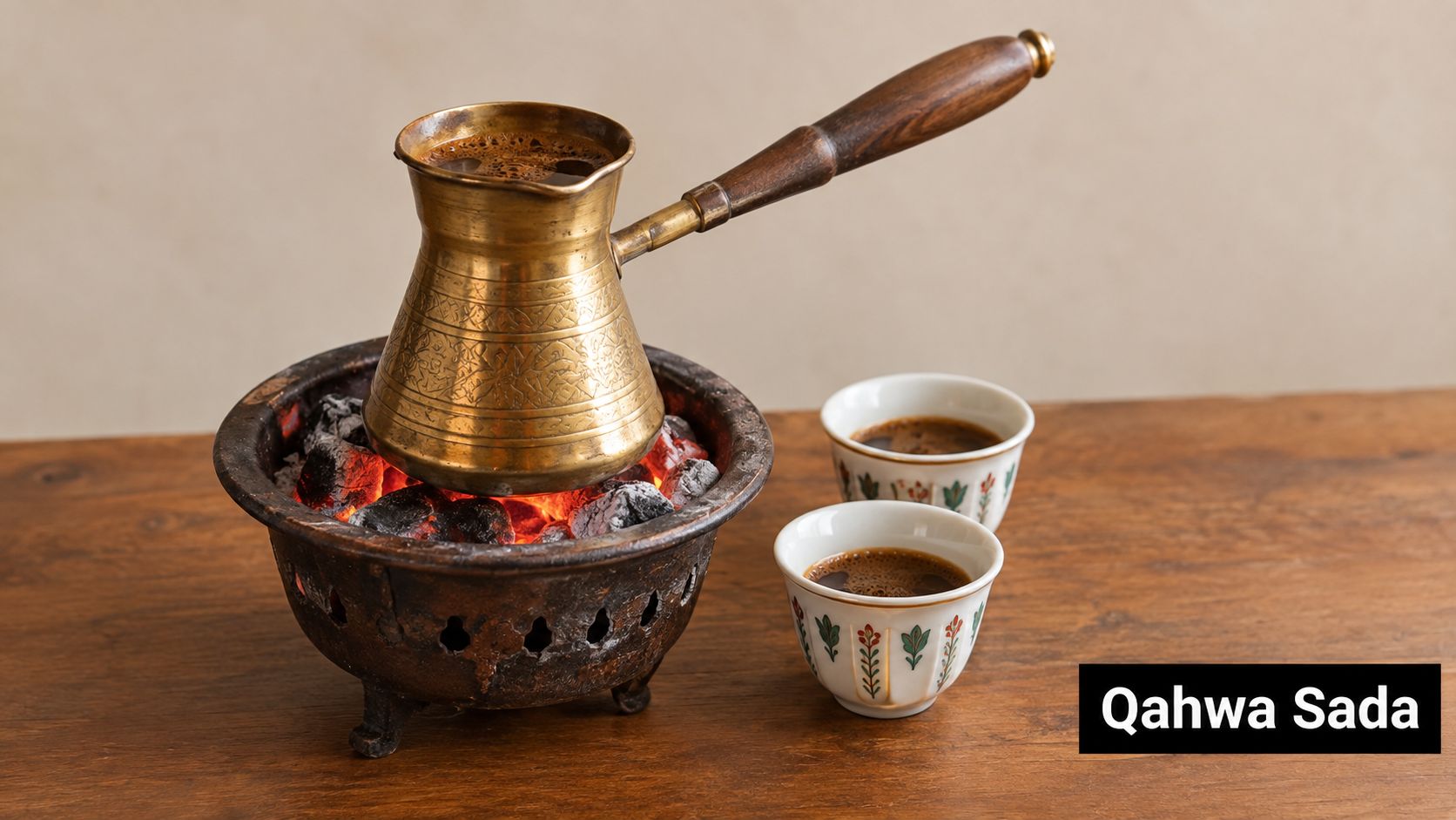 A traditional brass coffee pot sits on glowing coals next to two small decorative ceramic coffee cups.