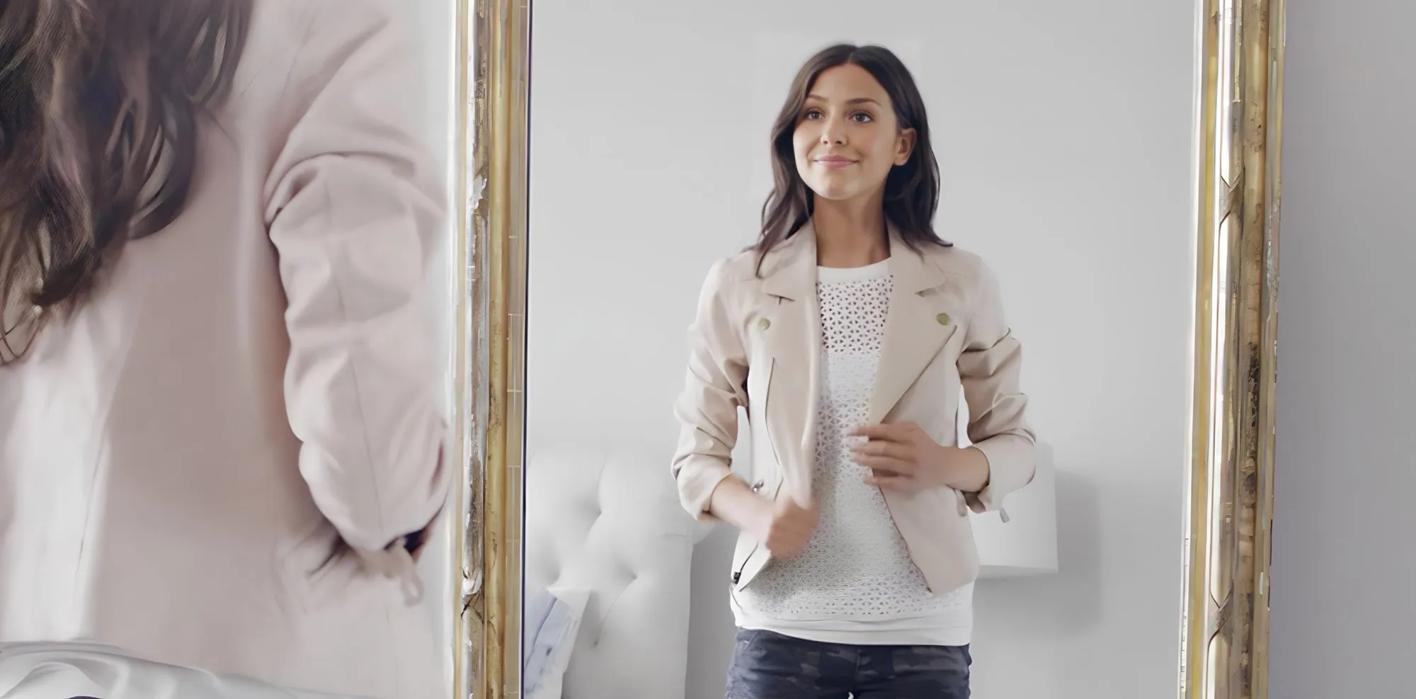 A woman smiles while looking at her reflection in a large mirror, wearing a light-colored jacket over a white patterned top.