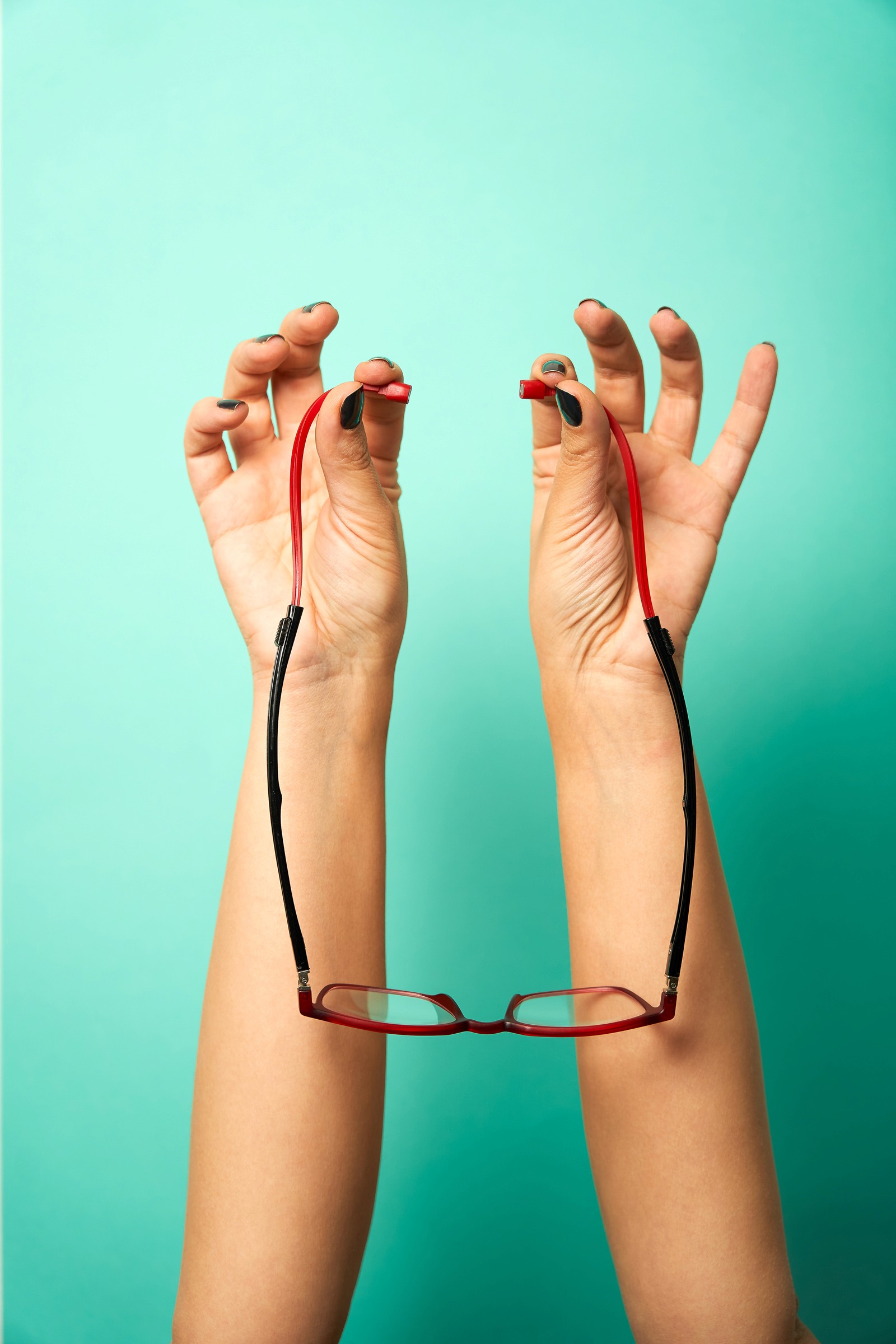 Photographie au flash sur un fond coloré vert d'une pair de mains tenant une paire de lunettes de lectures rouge et noir par ses branches flexibles 