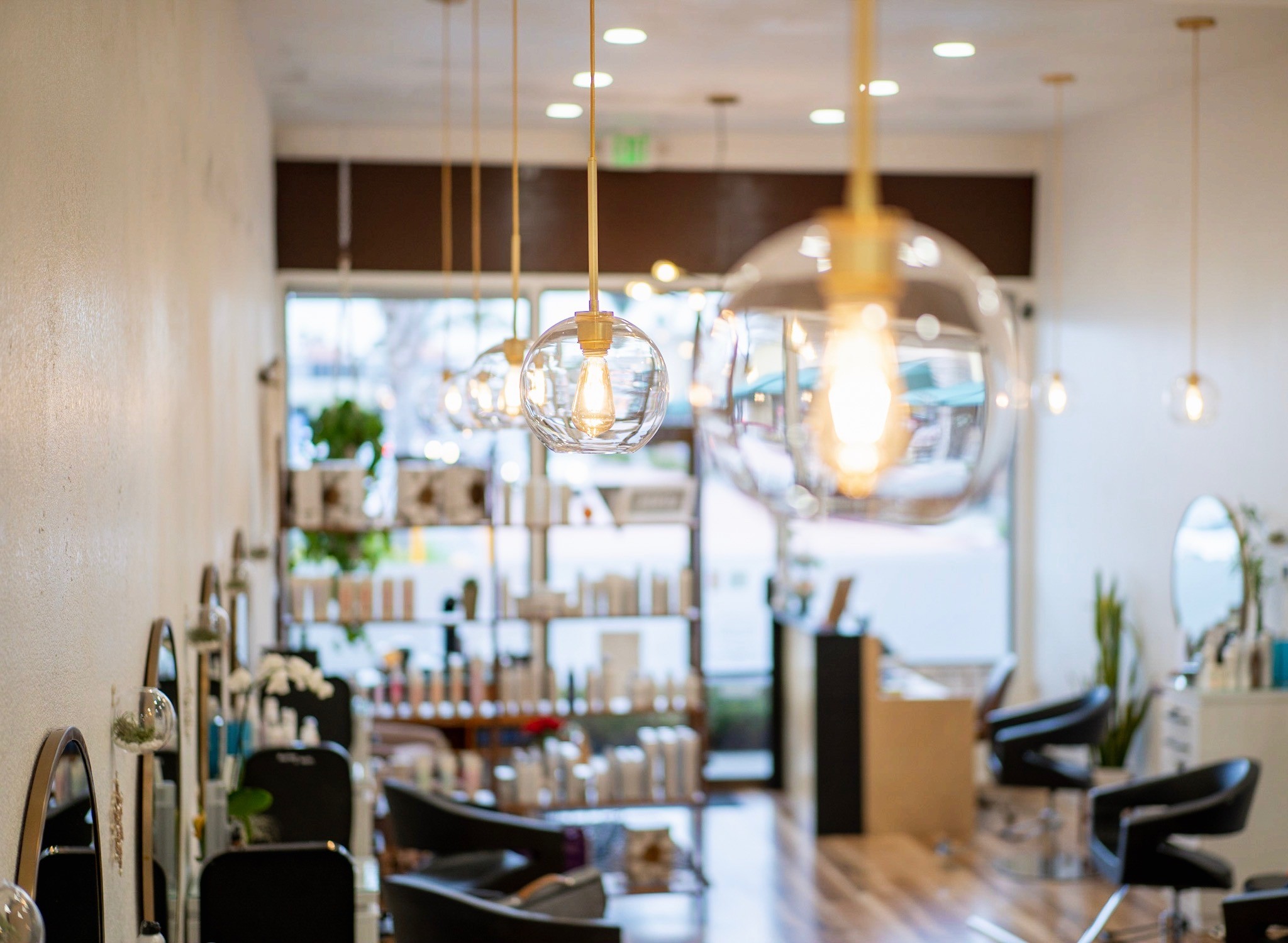 Wide-angle view of Del Sol Hair Studio, showcasing modern styling stations, soft pendant lighting, and retail shelves against natural light from the front windows.