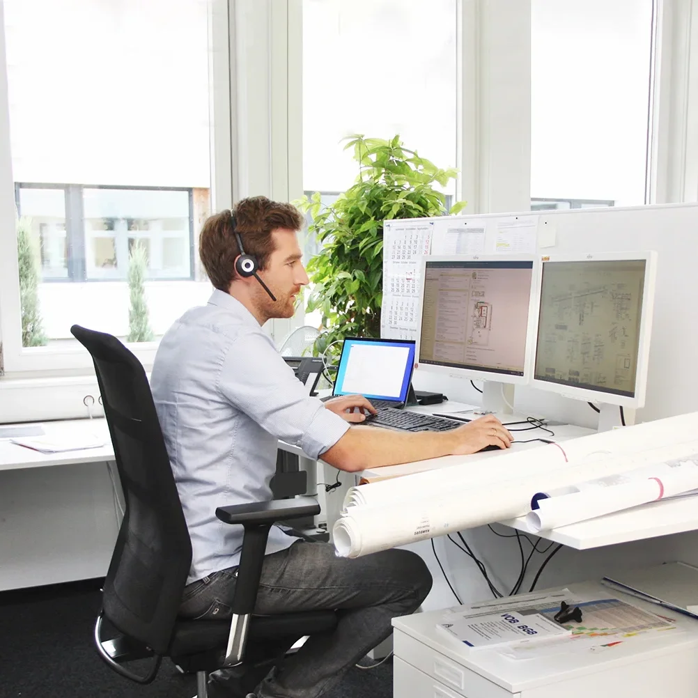 Mann mit Headset tippt auf Tastatur vor zwei Monitoren. Helliges Büro mit Pflanzen und gerollten Plänen.