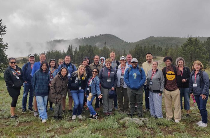 Group of around 25 people pose outdoors in front of misty pine-covered hills under a cloudy sky. Most wear jackets and conference lanyards, suggesting a professional outdoor excursion or retreat.v