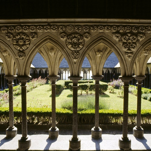 Ornate stone arches frame a garden courtyard with manicured bushes and flowers under a sunny sky.