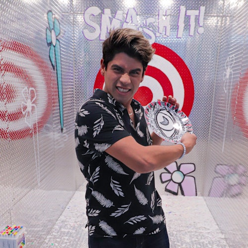 A person in a black patterned shirt holds a shattered plate and poses in front of a target with "SMASH IT!" written above.