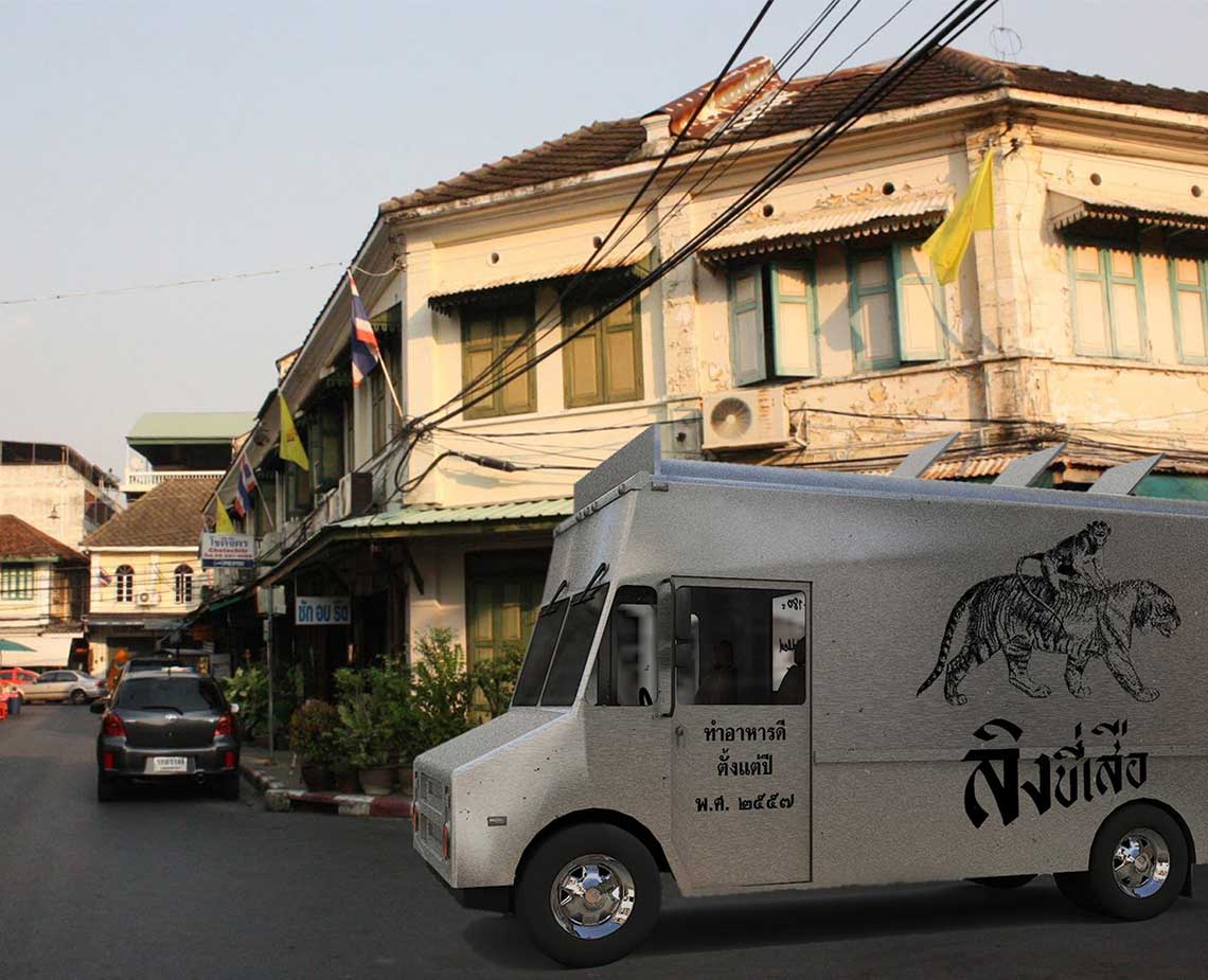 A white truck with a black tiger graphic and Thai script is parked on a street. The truck faces right, and its side displays Thai script. Behind it, a tan building with windows and balconies stands under a clear blue sky. A dark-colored car is parked to the left of the truck. Power lines run across the upper portion of the image.