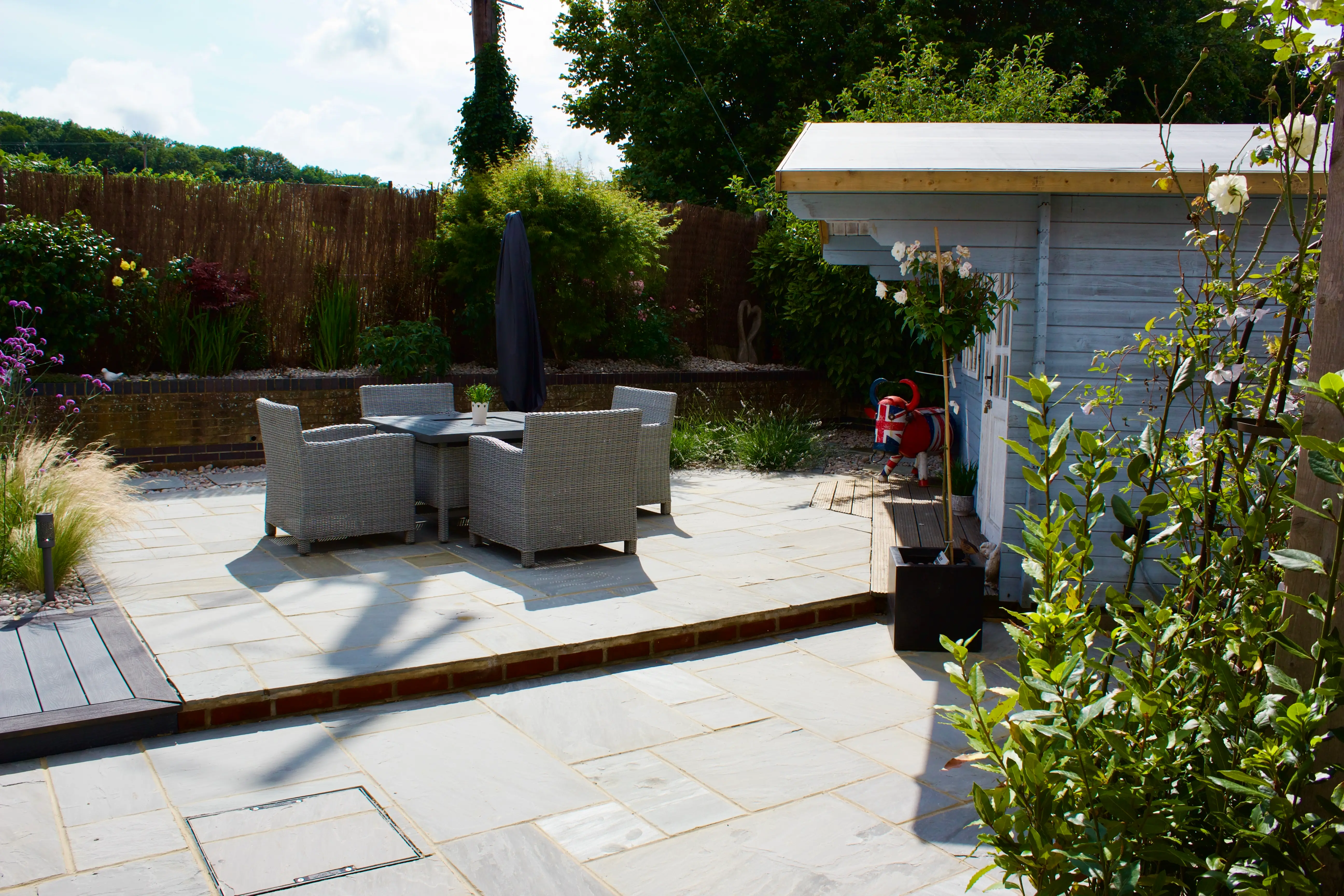 A patio area with stone tiles, a table and chairs, surrounded by plants and a shed in the background.