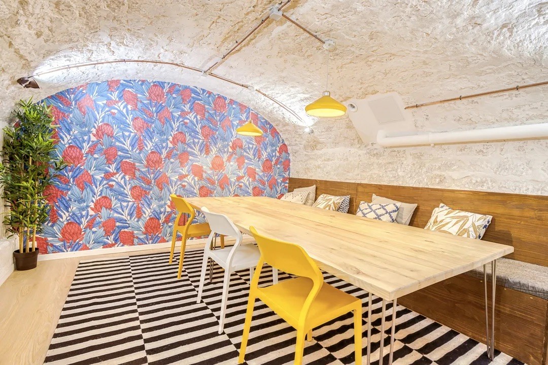 Coin convivialité dans un sous-sol voûté avec papier peint floral coloré, tapis graphique et table de réunion.