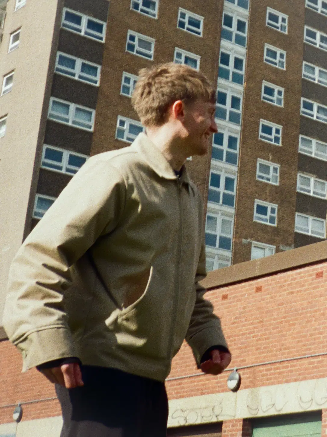 A person wearing a beige jacket is walking in front of a tall, brown brick apartment building with numerous windows, under a clear sky.