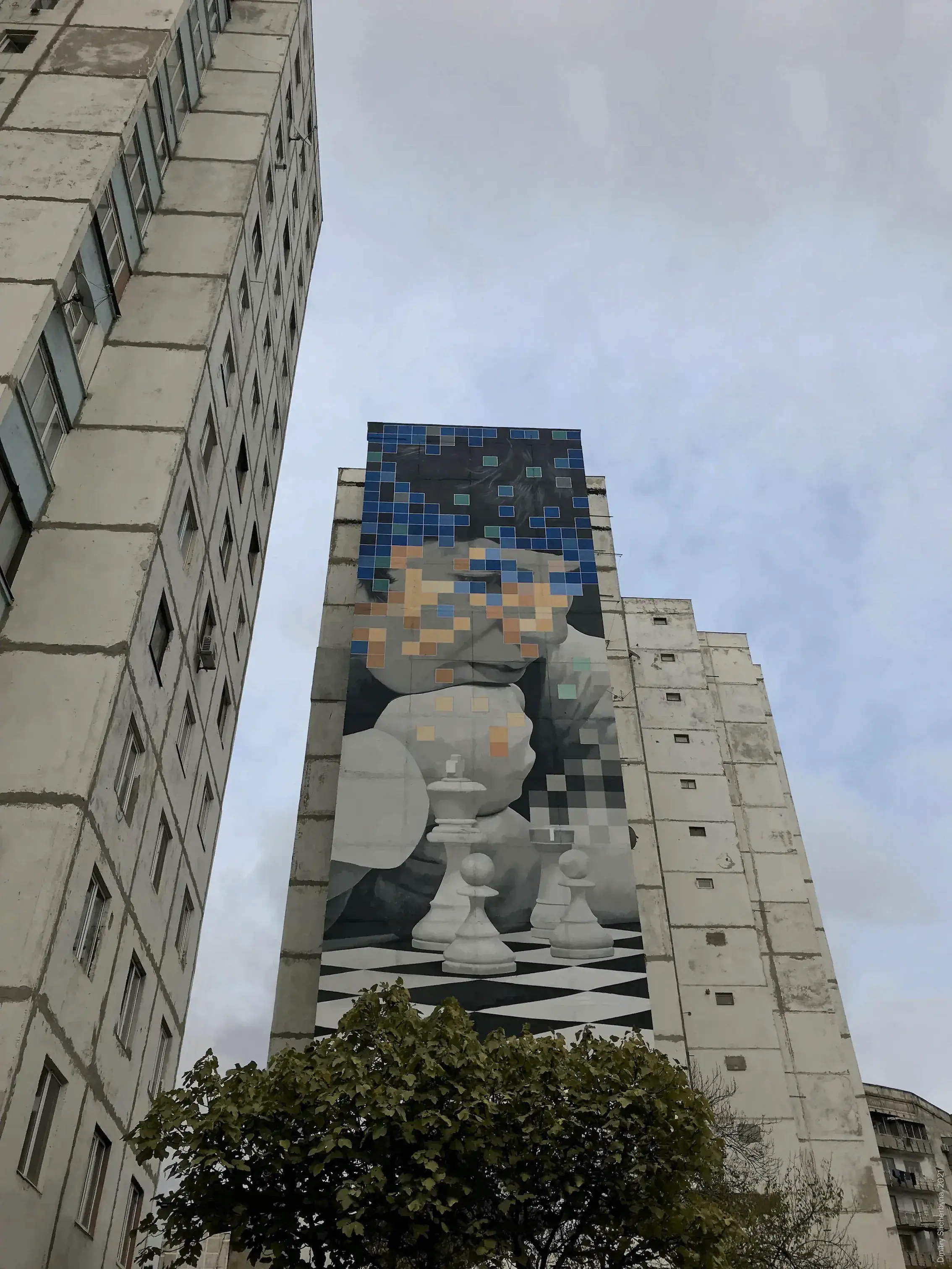 A tall mural on the side of a high-rise building depicts a figure leaning over a chessboard with white chess pieces. The figure's face dissolves into blue, teal, and tan pixel squares, blending realism with digital abstraction.