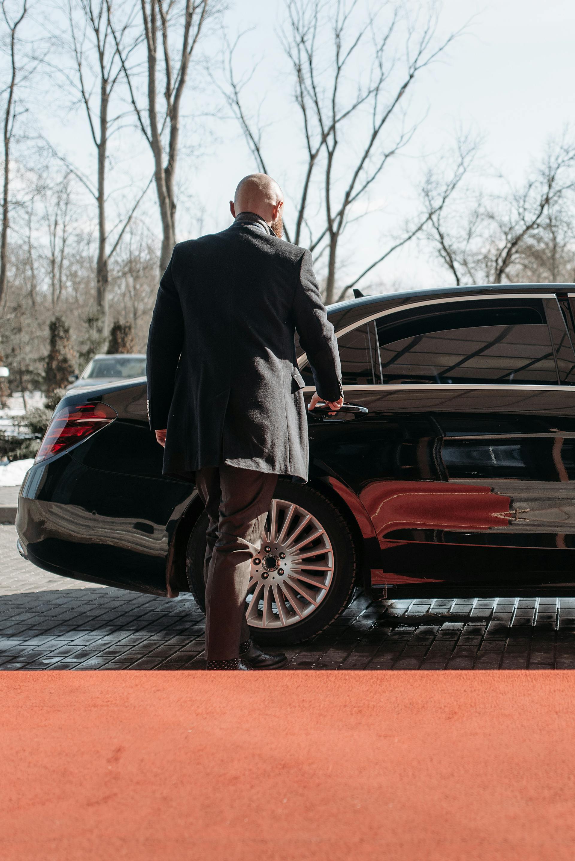 Luxury chauffeur opening the door of a black sedan on a red carpet, surrounded by trees, symbolizing premium airport transfer services.