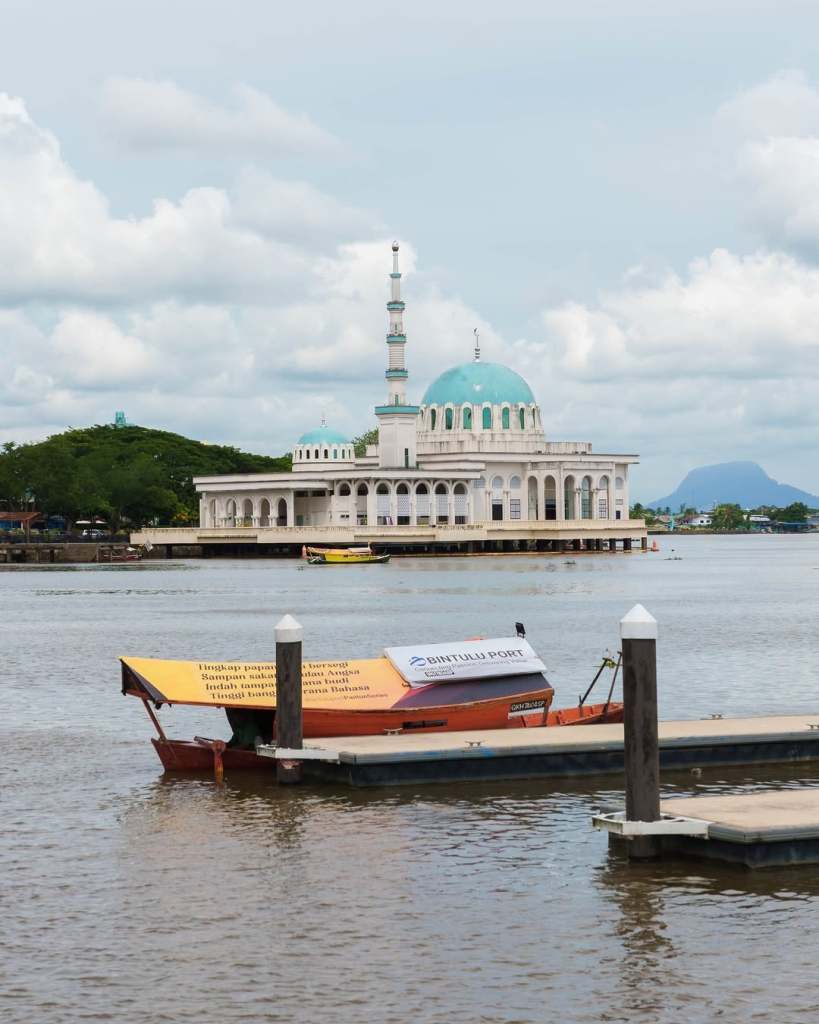 View of a mosque with turquoise dome and minaret on the riverside, surrounded by cloudy sky and boats anchored at a dock.
