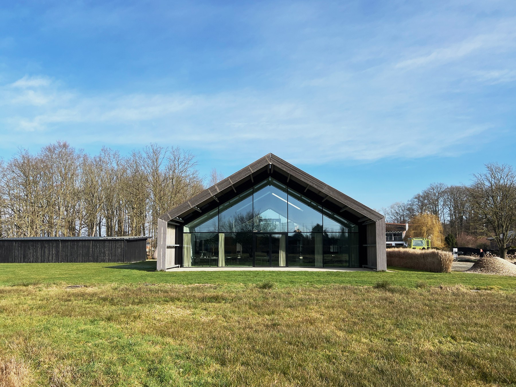 Vooraanzicht van een modern, schuur-achtig gebouw met een strak puntdak en een volledige glazen gevel. Het gebouw staat in een open grasveld onder een helderblauwe lucht, omringd door bomen zonder bladeren.