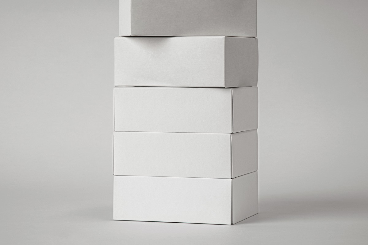 Stack of kraft cardboard boxes with bold black typography, arranged vertically against a dark background. Minimal lighting highlights the texture and structure of the packaging.