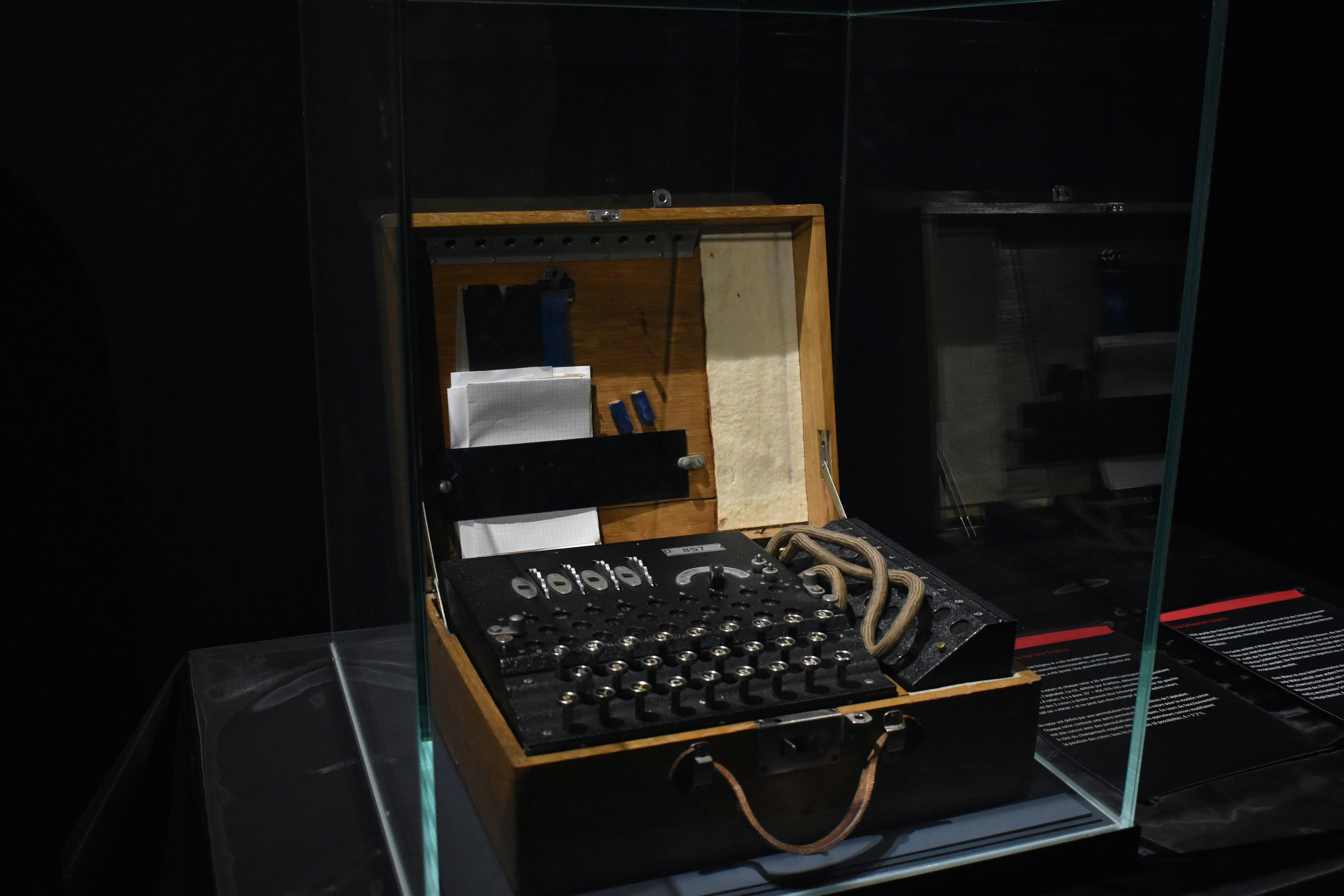 A vintage Enigma machine in a wooden carrying case, displayed in a glass museum case. The black typewriter-like device features rows of keys and is housed in a hinged wooden box. The machine appears to be in a dimly lit exhibition space.