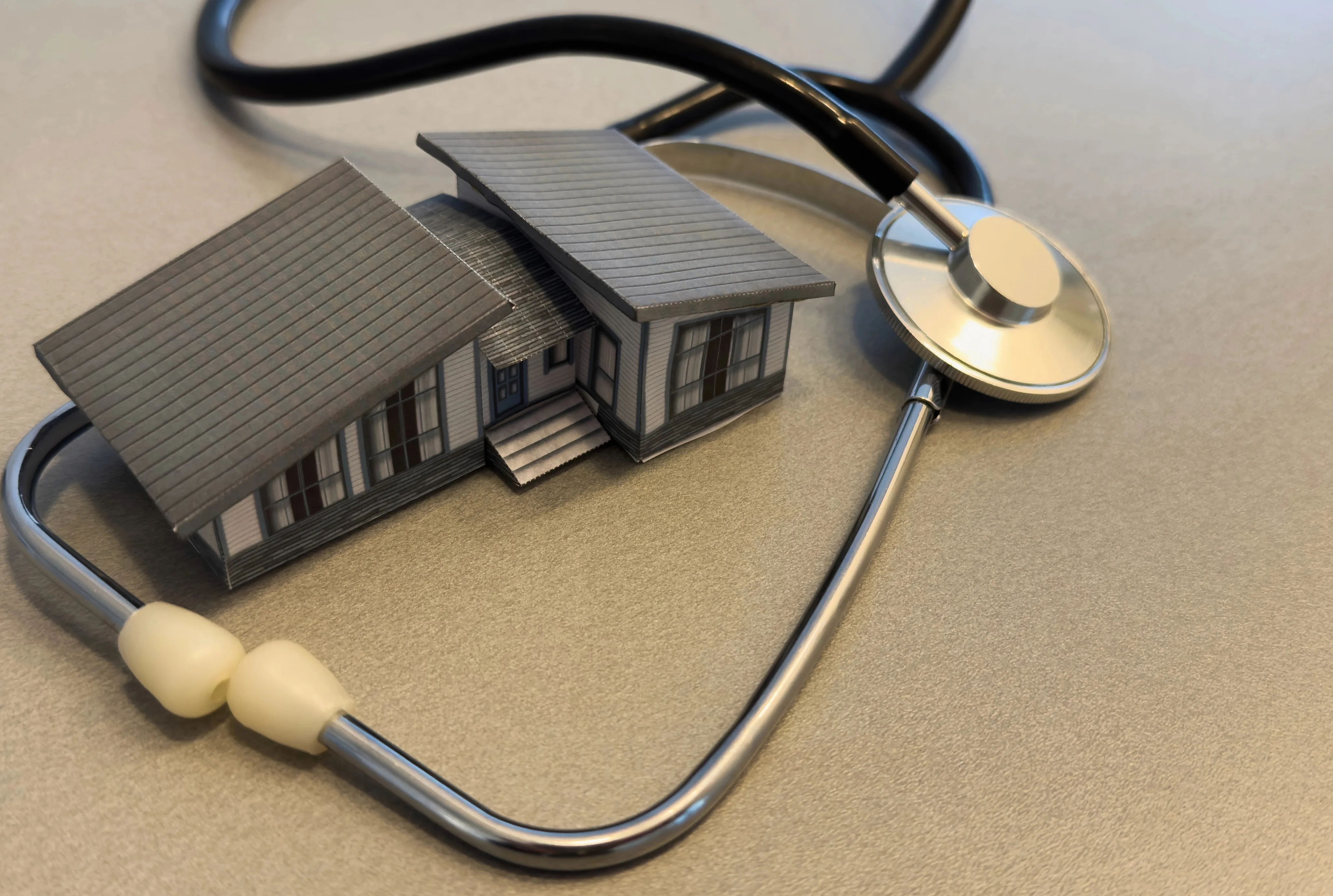 A stethoscope coiled around a miniature gray house model on a beige surface, symbolizing home health care and medical property inspection.