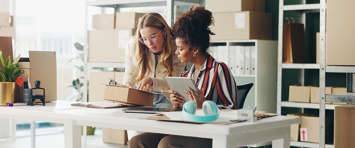 Two women sitting at a kitchen table, looking at a laptop and smiling, with a few plants in the background.