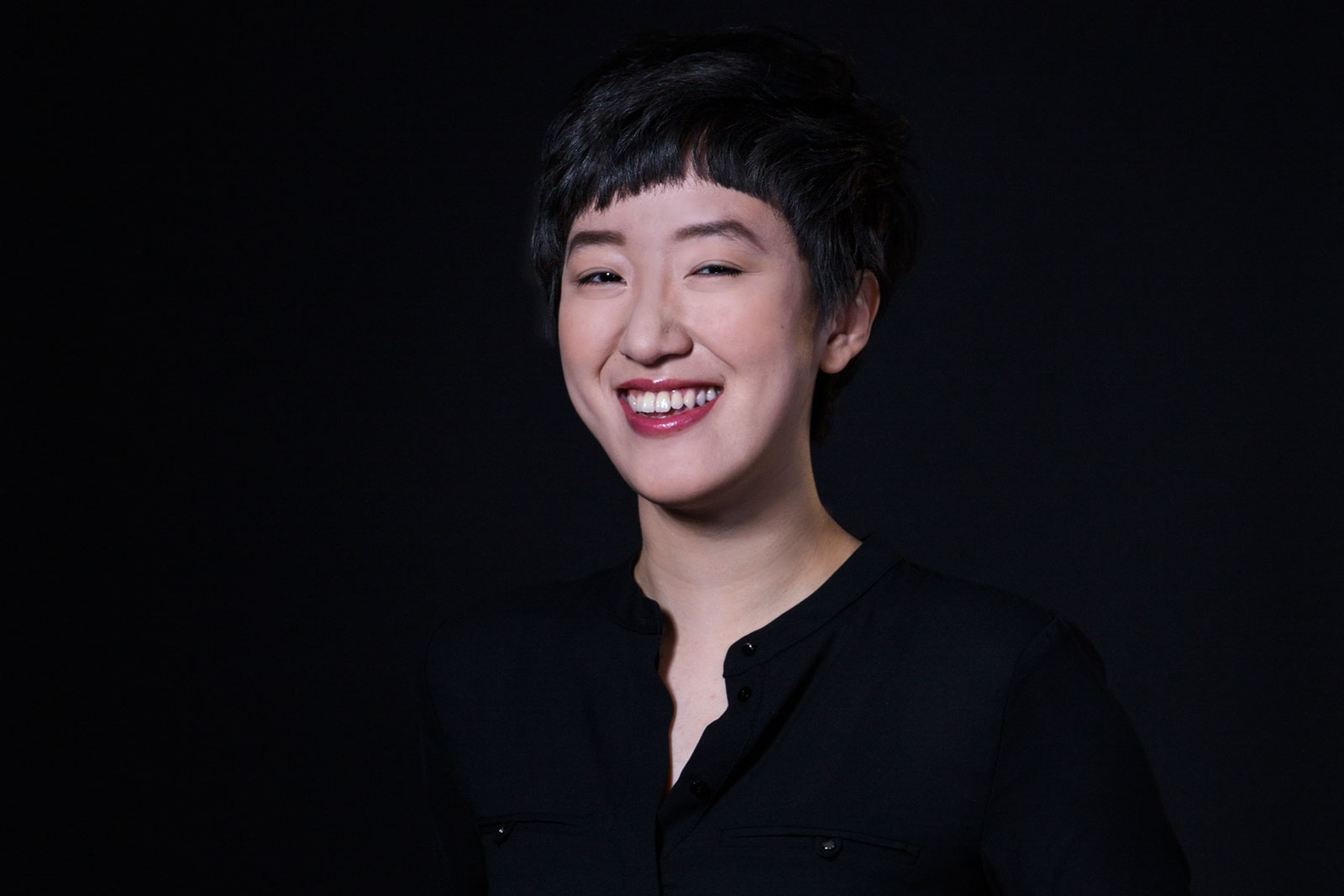 A person with short, dark hair and dark eyes is smiling, wearing a black button-up shirt against a solid black background. Their head is slightly tilted to the left, and their mouth is open, revealing teeth. The lighting highlights their face and the top of their shirt.