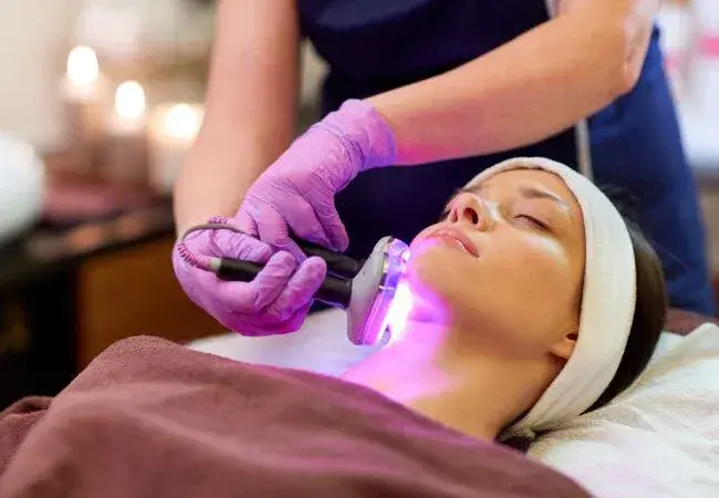 Woman receiving light therapy facial treatment using LED and IPL technology to reduce acne, improve skin tone, and stimulate collagen production.