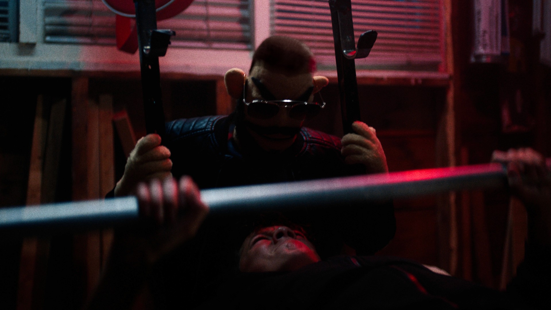 A puppet with sunglasses holds a weight bar over a young boy's face, creating a tense atmosphere. The setting is dimly lit with an ominous red glow.