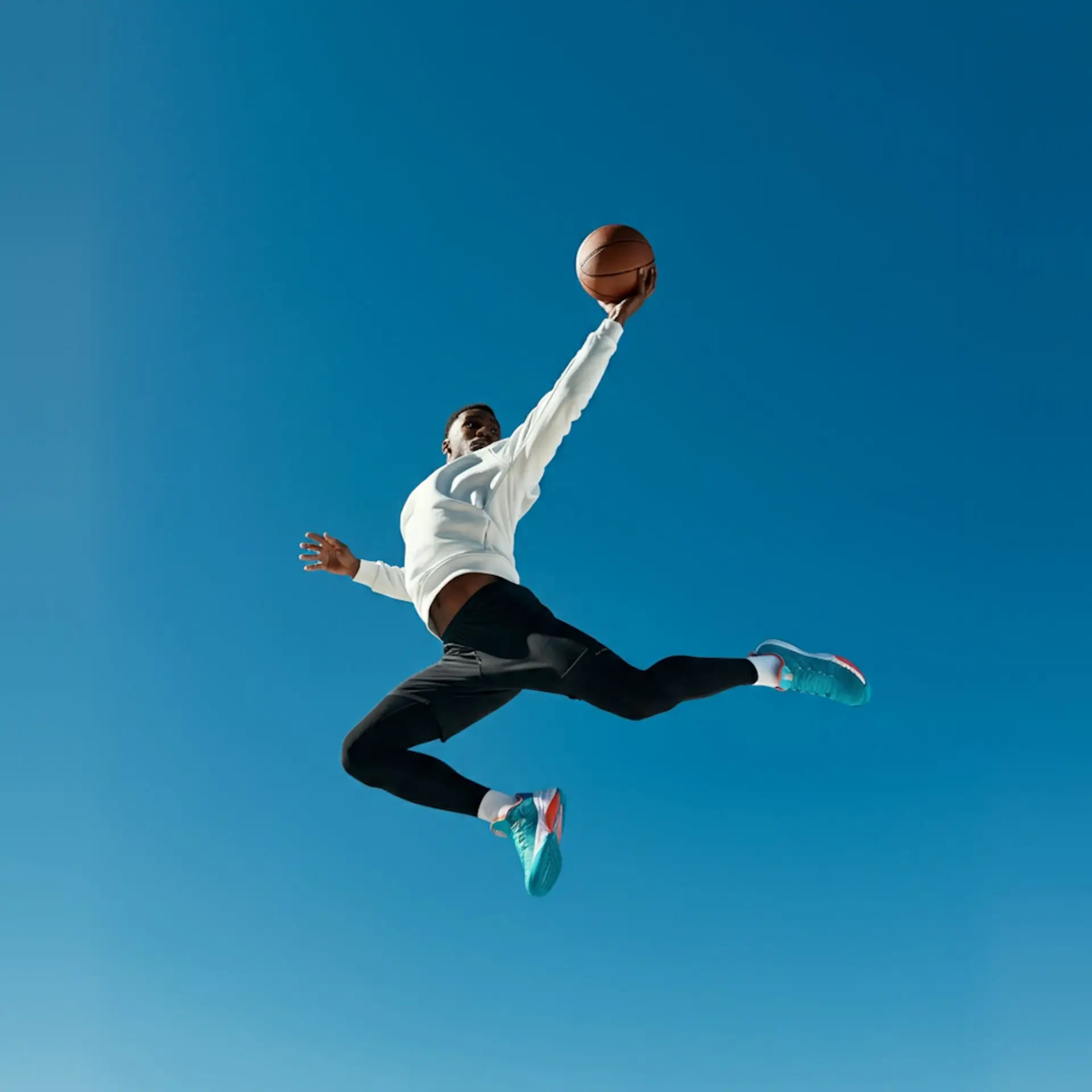 A basketball player in mid-air against a clear blue sky, reaching for a dunk.