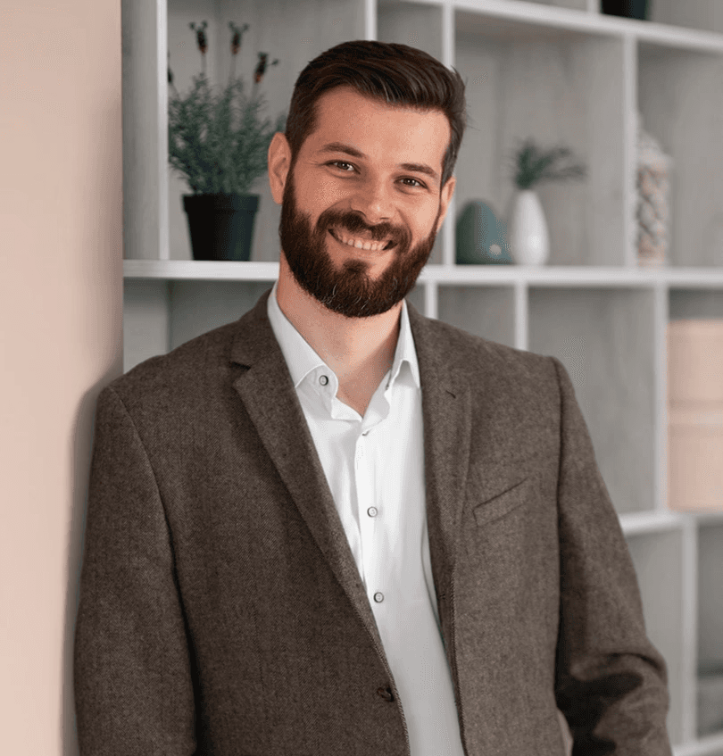 Smiling man with a beard, in a brown jacket, leans against a wall holding a laptop. Behind him is a shelf with plants and decorative items.