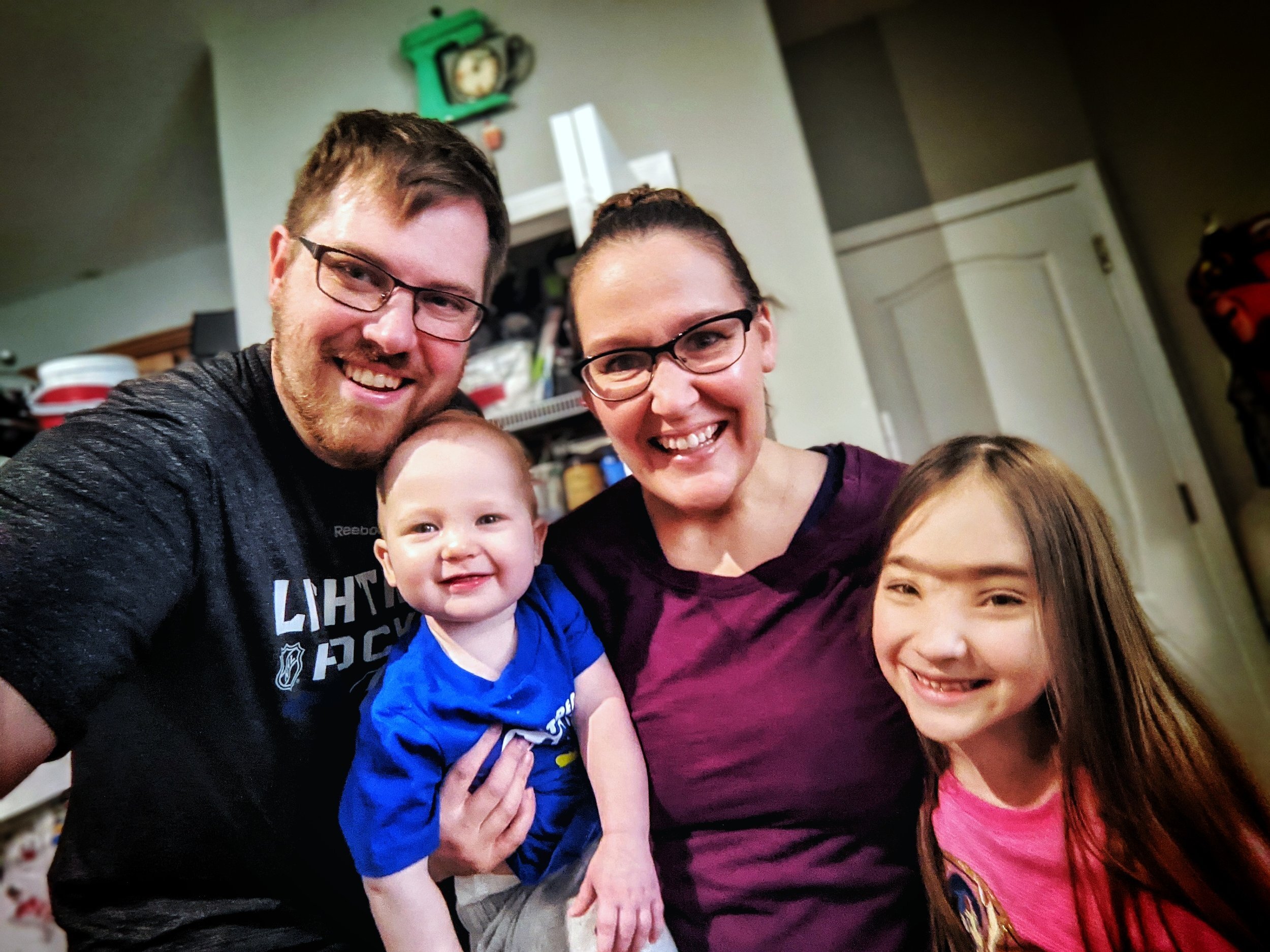 Alt text: Smiling family of four taking a casual indoor selfie, with two adults and two children posing closely together in a kitchen setting.