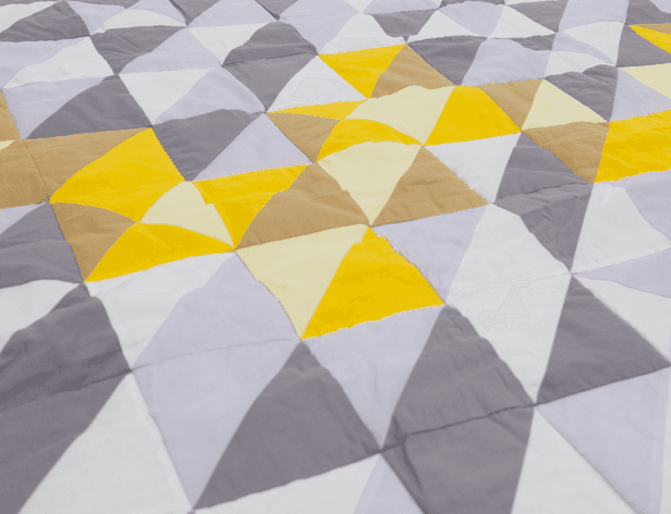 A close-up view of a quilt or blanket with a geometric pattern of triangles in yellow, gray, and off-white. The triangles are arranged to create a repeating, symmetrical design. The quilt is folded or draped, with some areas showing slight wrinkles or creases.