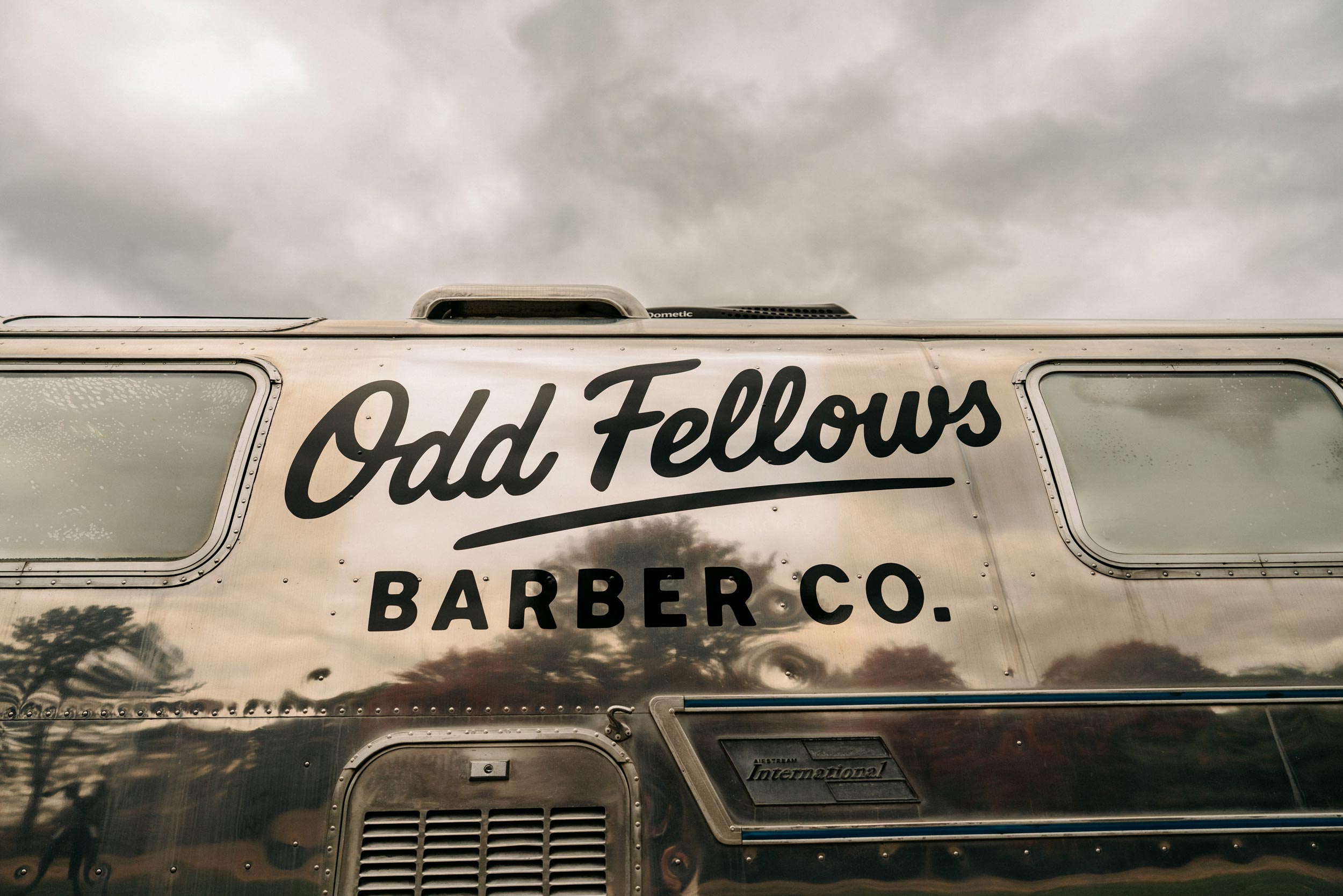 A close-up shot of the side of a shiny, silver Airstream-style trailer with the words "Odd Fellows BARBER CO." painted in black. The reflection of the surroundings is visible on the highly polished surface, and the sky above is mostly cloudy.