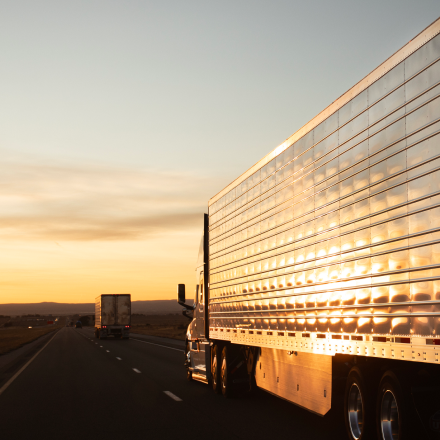 Refrigerated semi-truck trailer traveling on a highway at sunset, reflecting golden light off its polished aluminum sides.