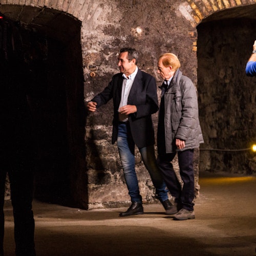两名男子走在一个光线昏暗、拱形天花板的石制走廊中；一位穿着西装外套和牛仔裤，另一位穿着灰色外套。