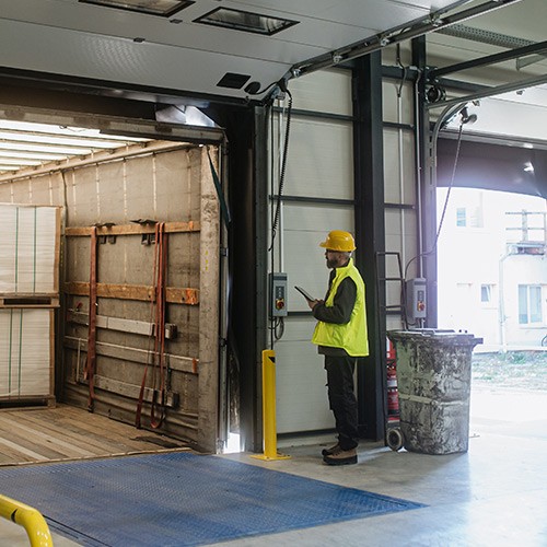 Photo of warehouse loading dock with door open. Man in yellow vest looking at shipping contents.
