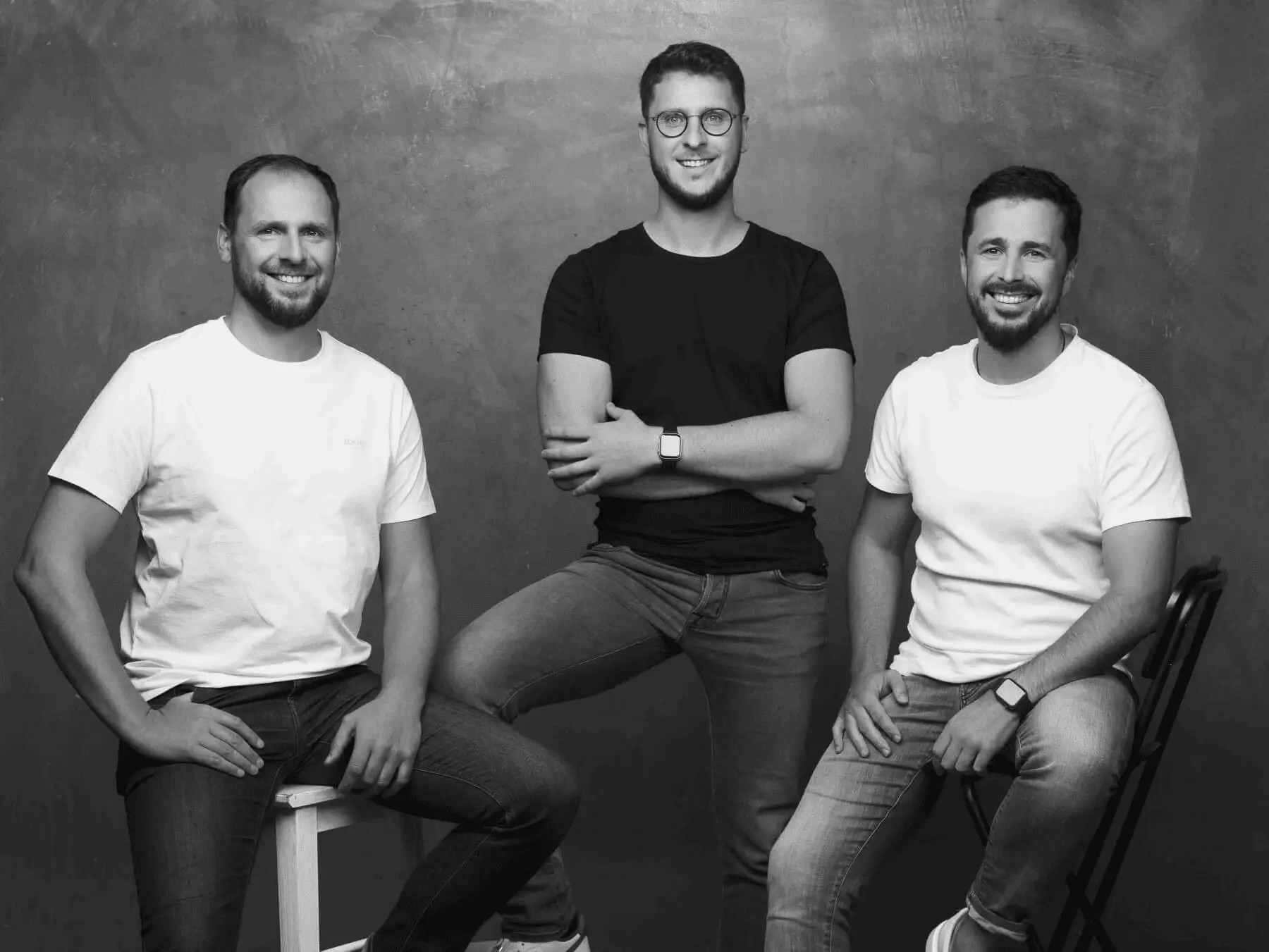 A black and white group portrait of three smiling men standing together against a dark background.