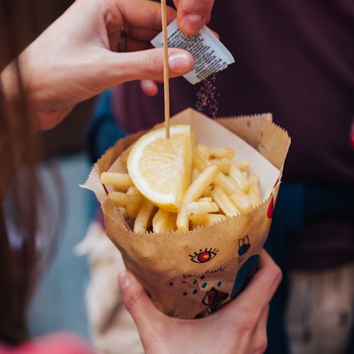 Osoba posypává kořením porci hranolků, které jsou ozdobeny citronovým klínkem, pomocí malého dřevěného špejle.