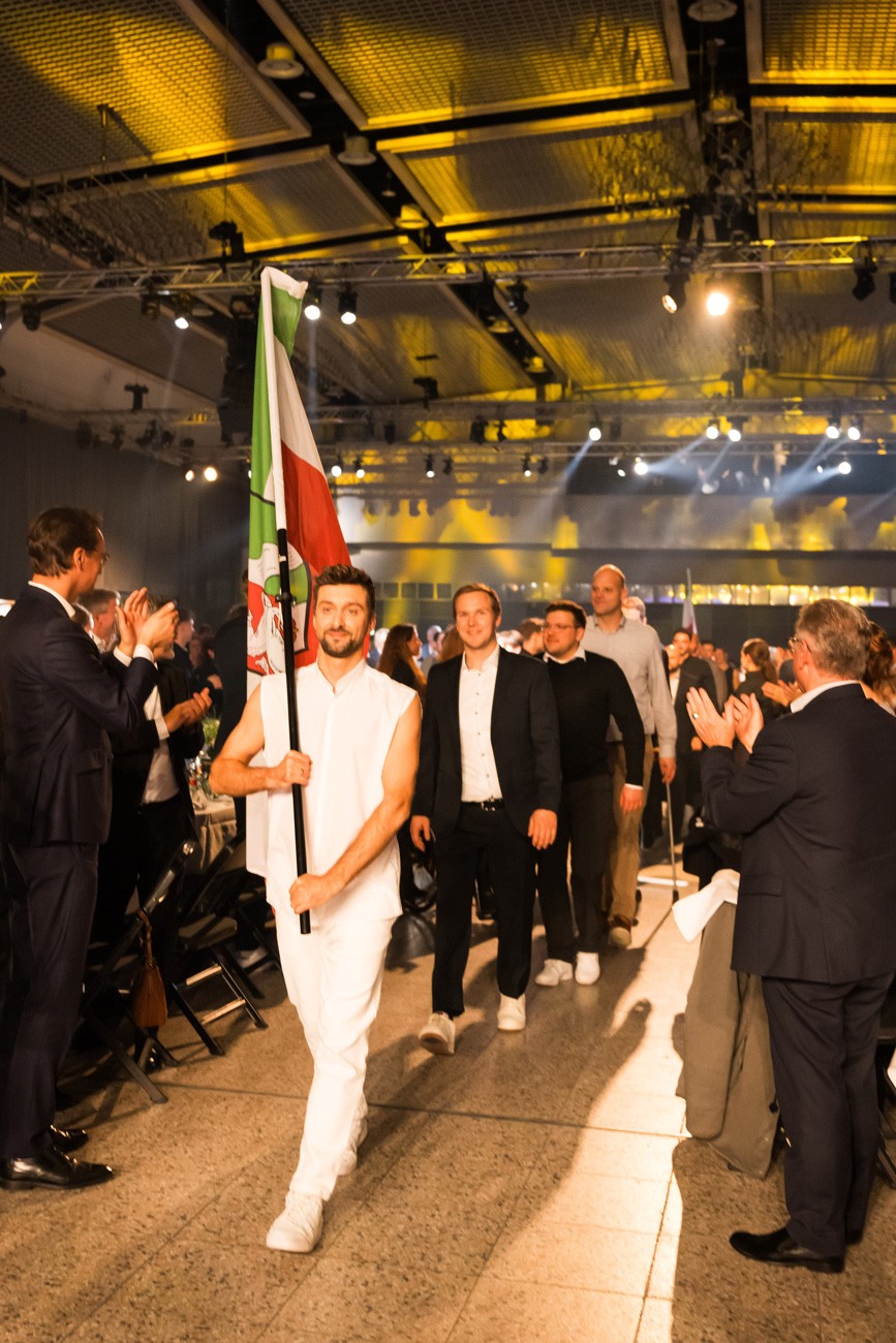 Mann mit weißer Kleidung trägt NRW-Fahne geht durch das Publikum und Gäste klatschen