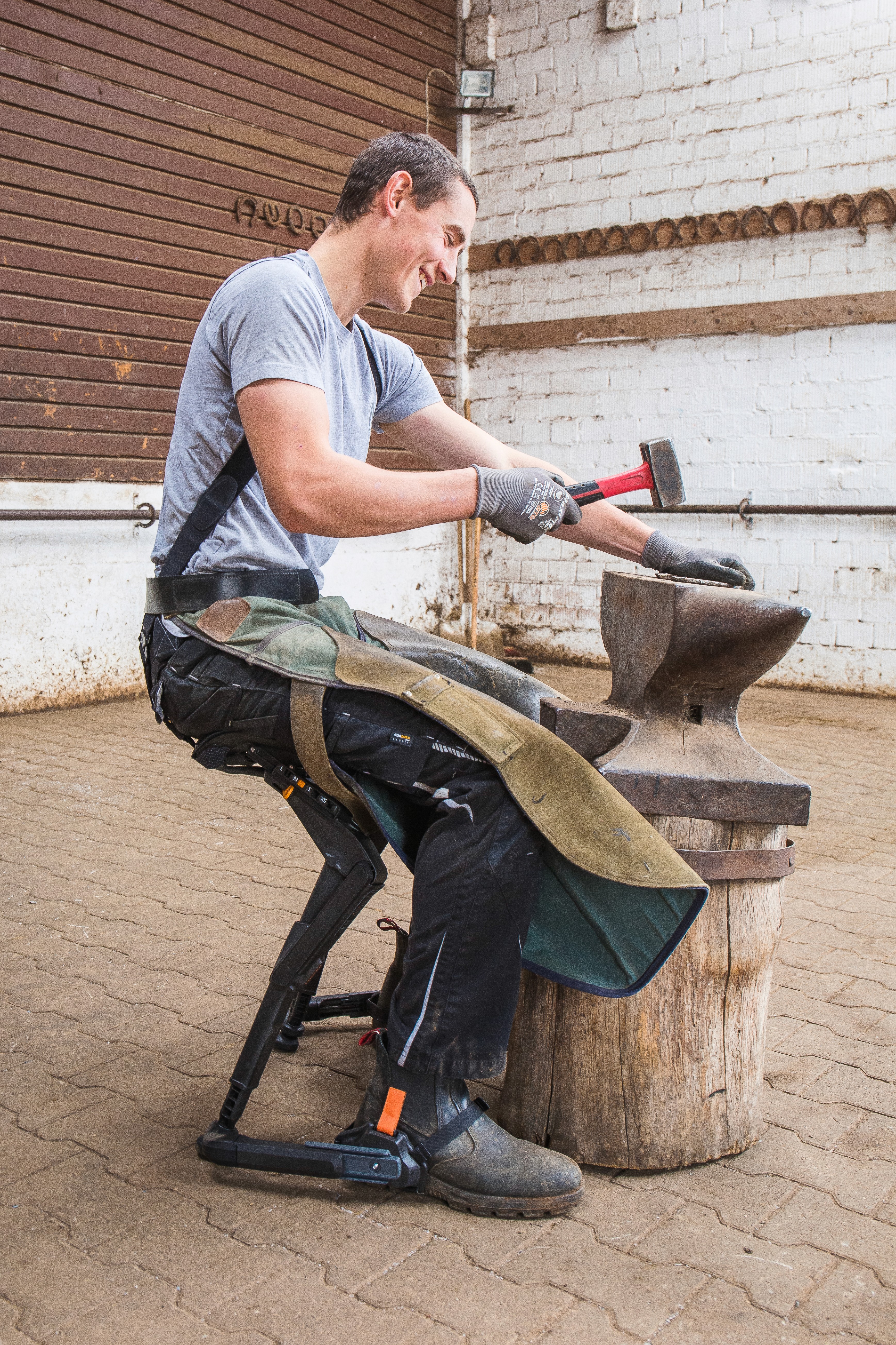 Travailleur assis sur un exosquelette de soutien des jambes de type chaise portative, utilisant un marteau sur une enclume dans un atelier, démontrant la réduction de fatigue lors du travail en position semi-accroupie.