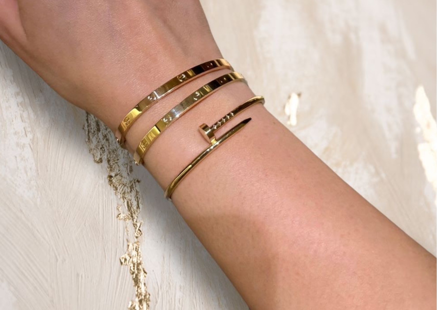 Arm adorned with gold and rose gold luxury, designer, branded, elegant, chic, classy, stylish Cartier jewellery in Love and Juste un Clou bracelets, shining in the light - against a white and gold marble background.