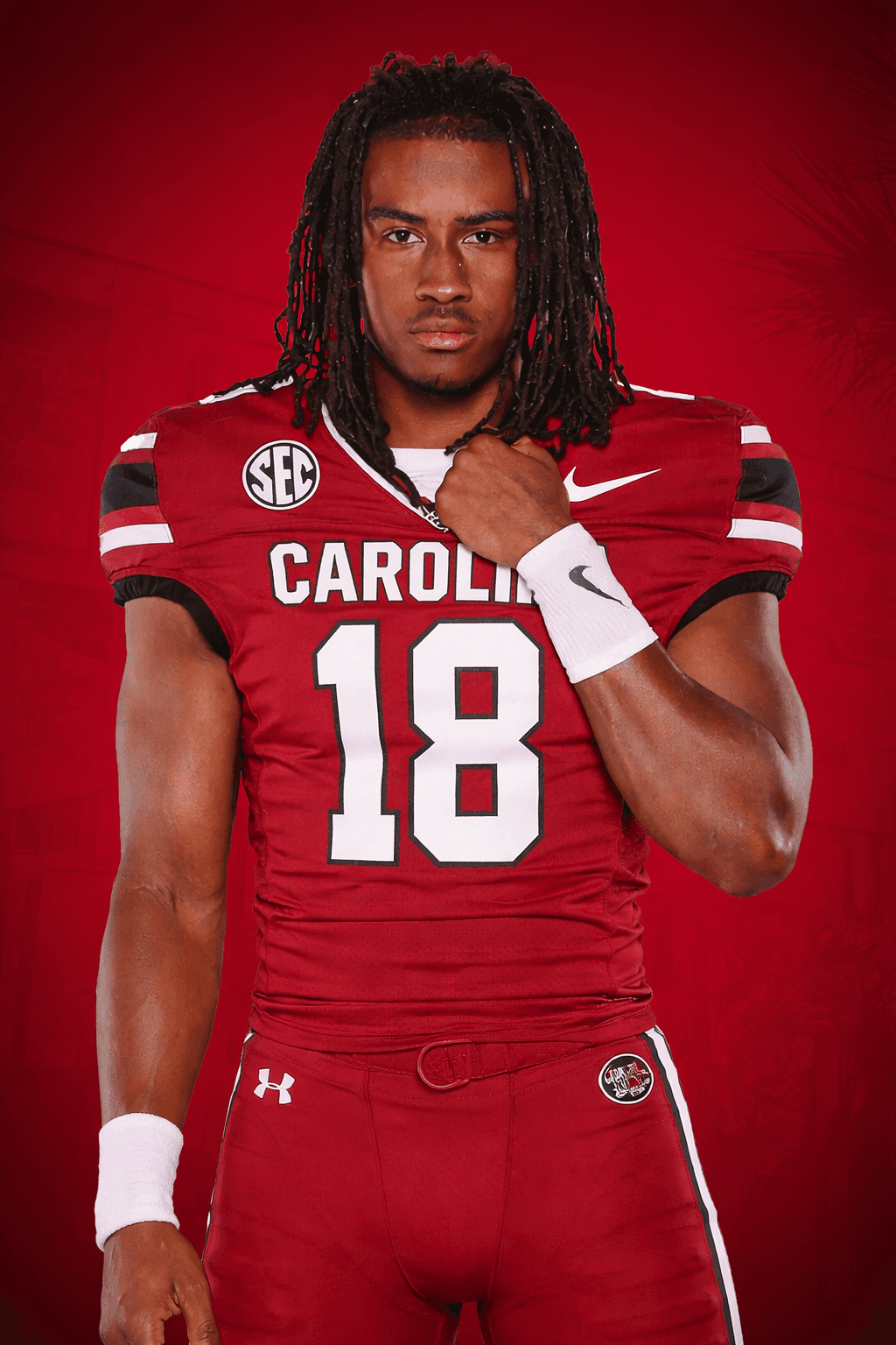 Lucian Anderson wearing a red South Carolina Gamecocks uniform with the number 18 stands against a red background, looking at the camera and holding his collar with one hand.