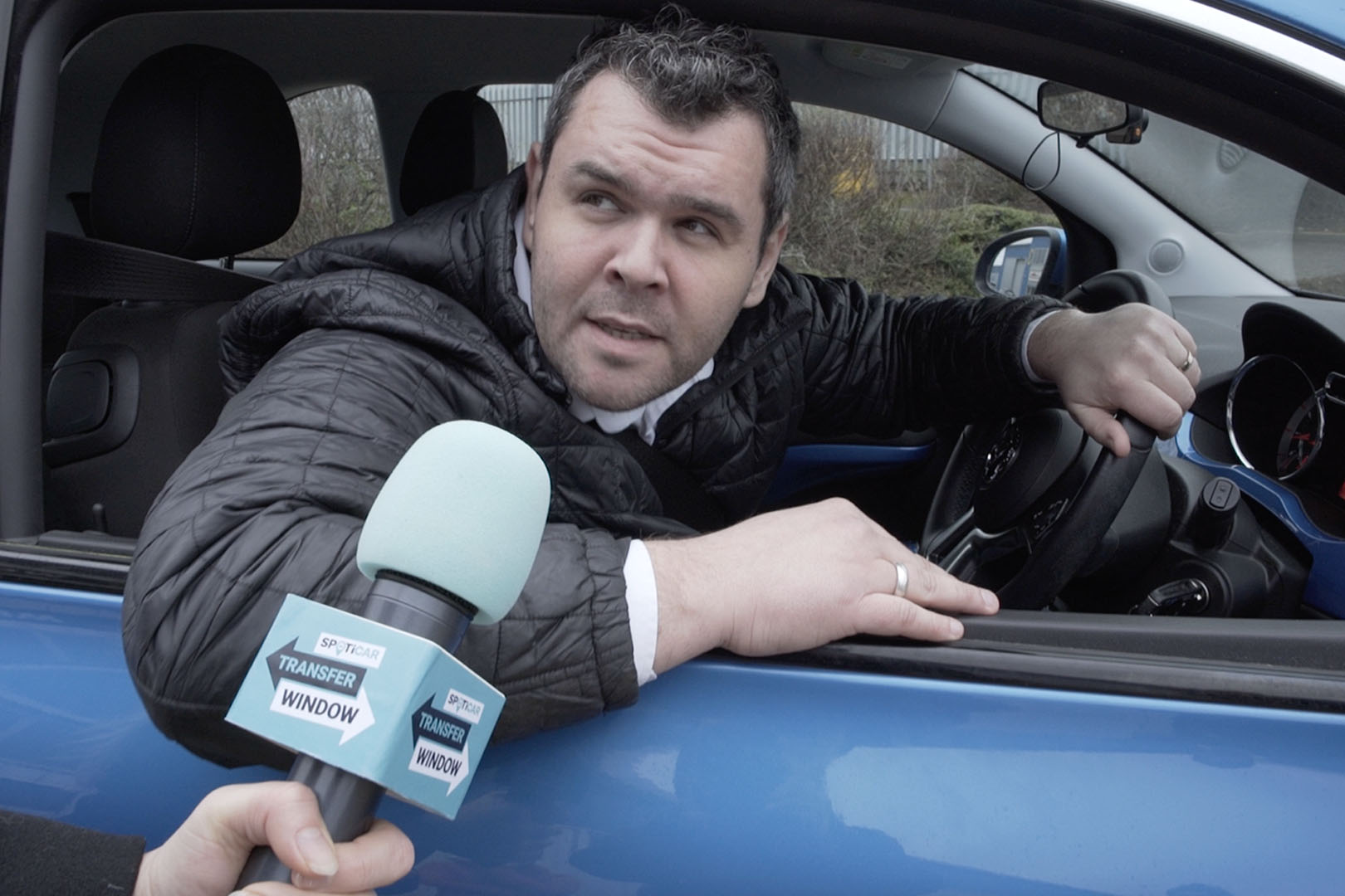 A man leaning out a car window, someone is holding a microphone towards him